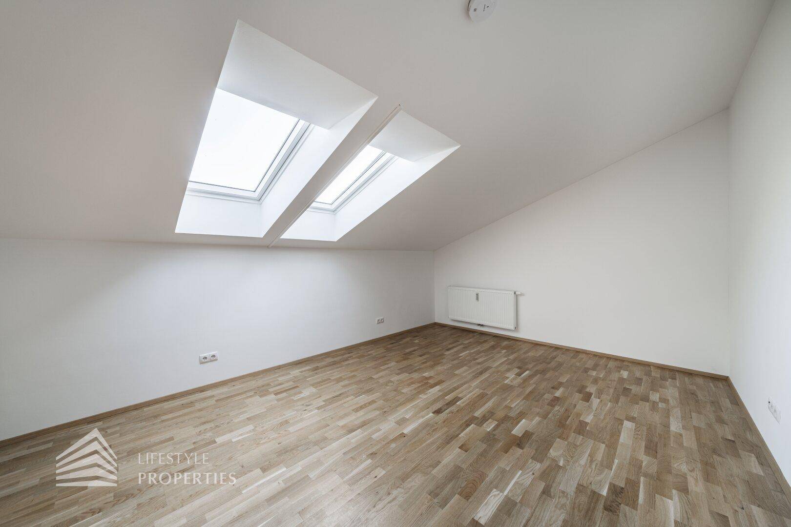 Geräumiges Zimmer im Dachgeschoss mit schrägen Wänden und zwei Dachfenstern.