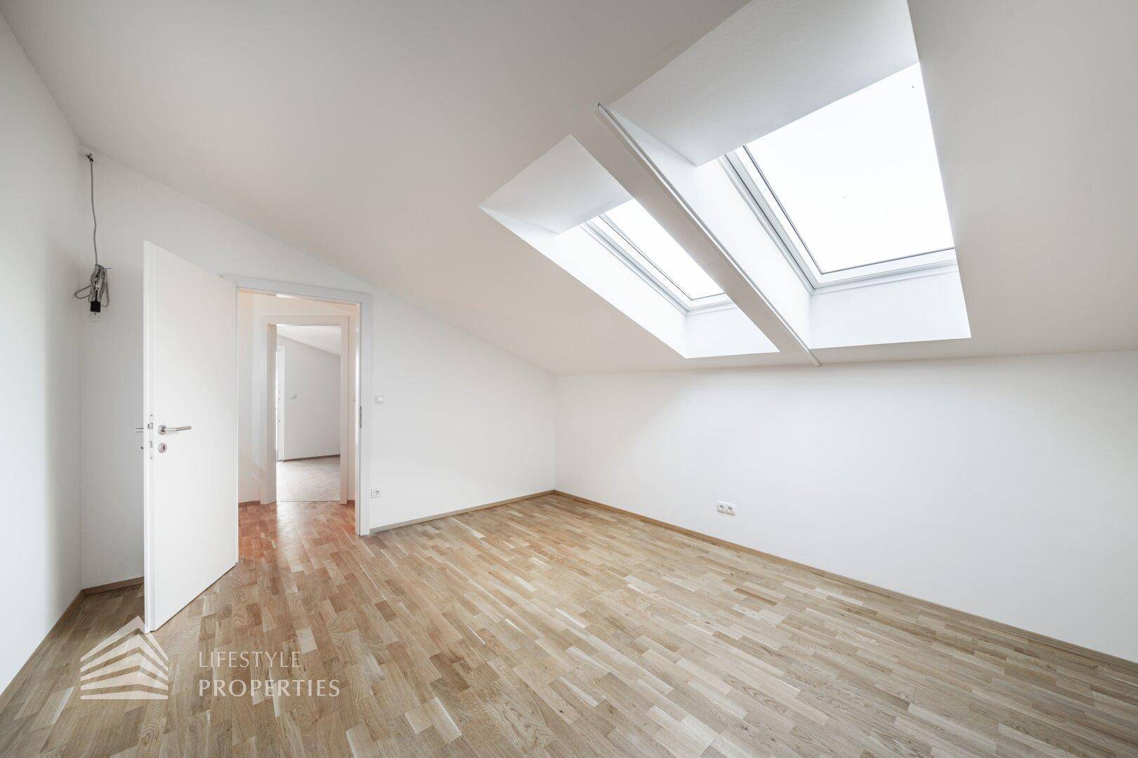 Helles Dachzimmer mit Holzboden, schrägen Wänden und zwei großen Dachfenstern.