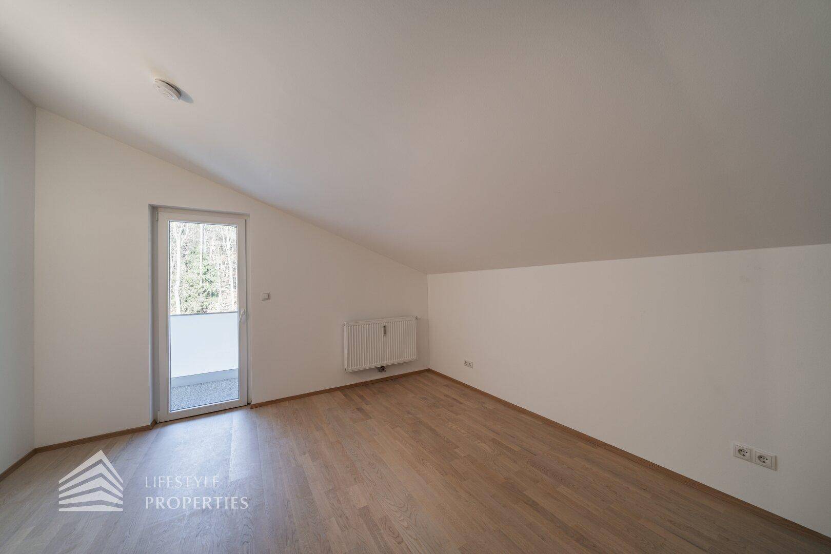 Dachgeschosszimmer mit Holzboden, schrägen Wänden und Zugang zu einem Balkon.