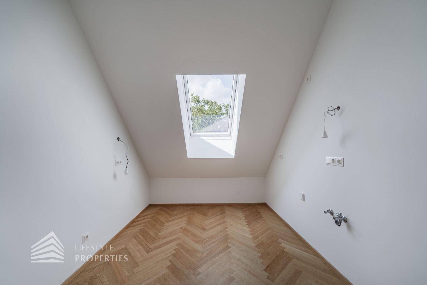 Helles Dachgeschosszimmer mit schrägen Wänden, Fischgrätparkett und einem großen Dachfenster.