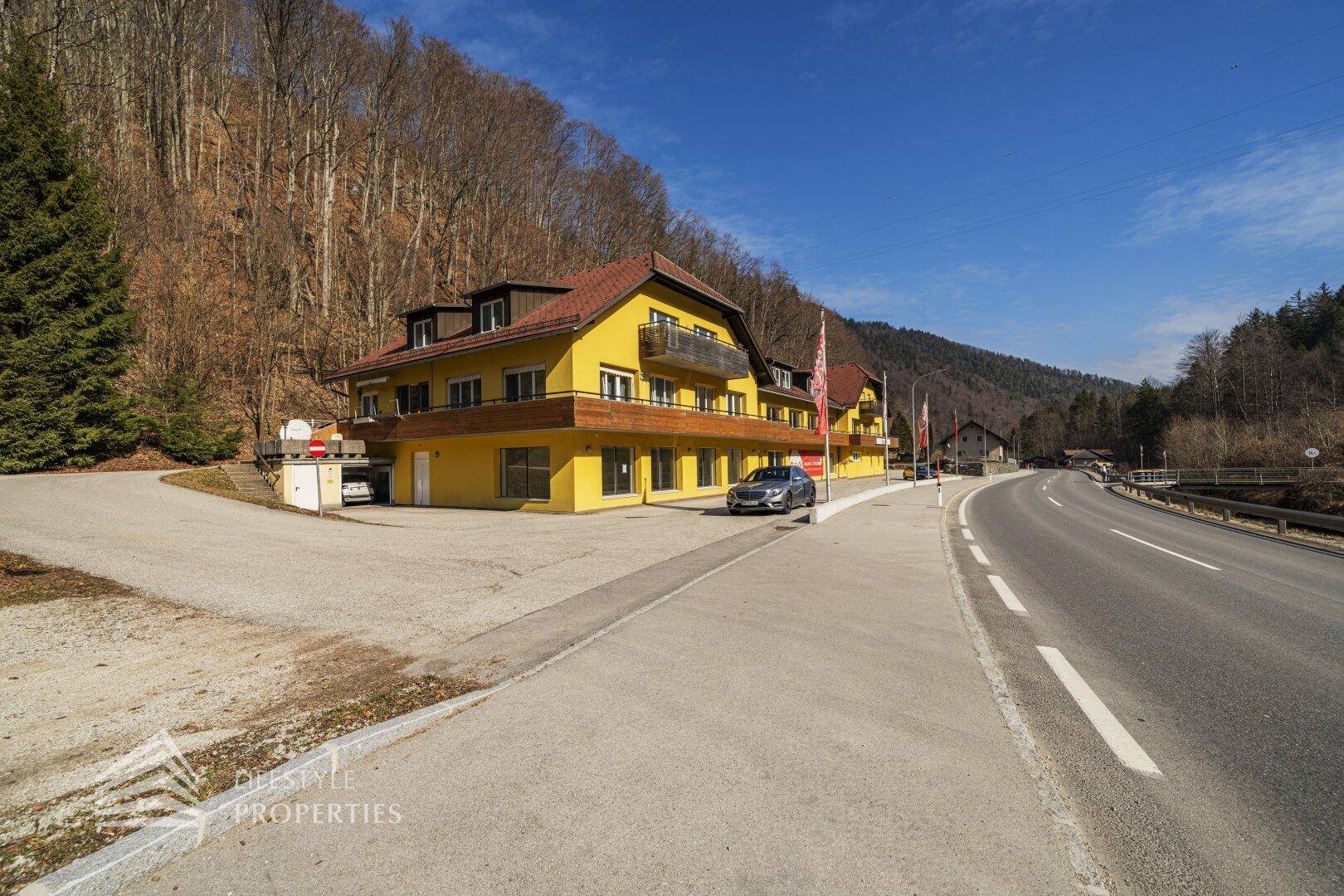 Gesamtansicht des gelben Gebäudes mit Zufahrt, Parkplätzen und umgebender Naturlandschaft.