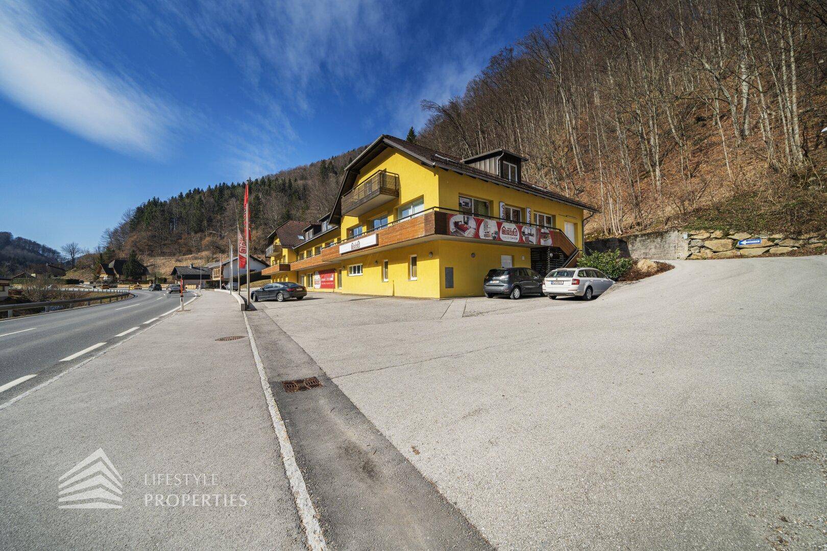 Ansicht des Gebäudes von der Straße mit Parkplätzen und der umliegenden bergigen Landschaft.