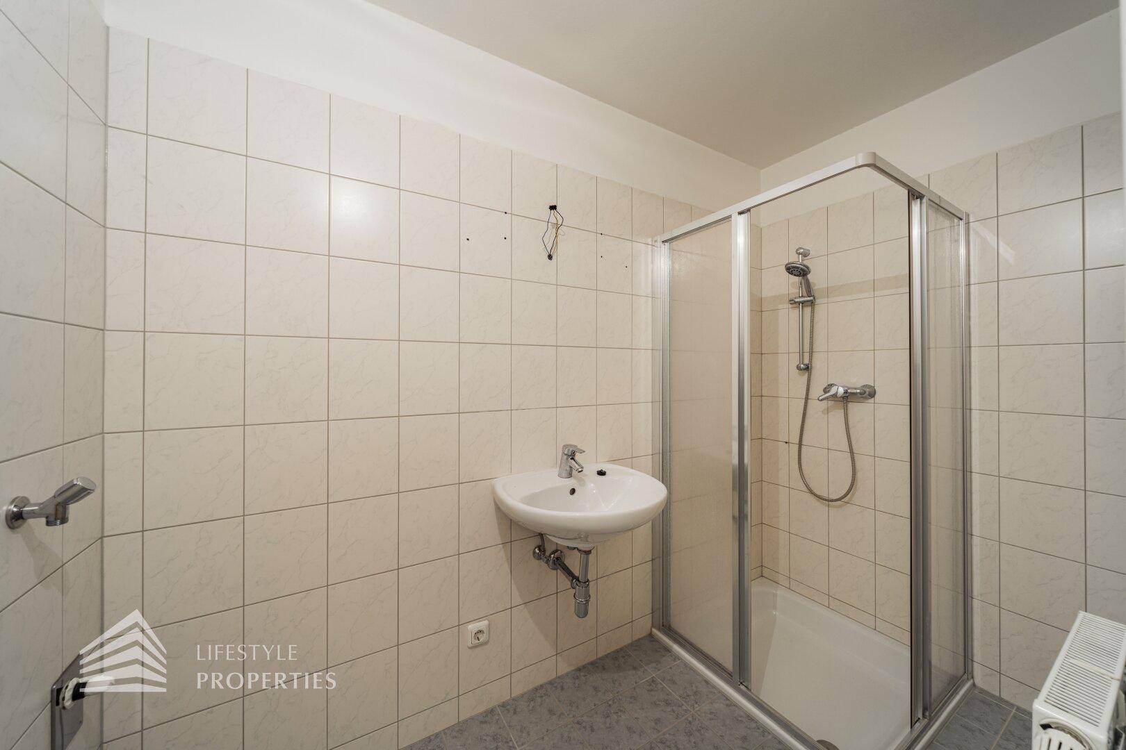Funktionales Badezimmer mit Duschkabine, Waschbecken und hellen Fliesen an Wänden und Boden.