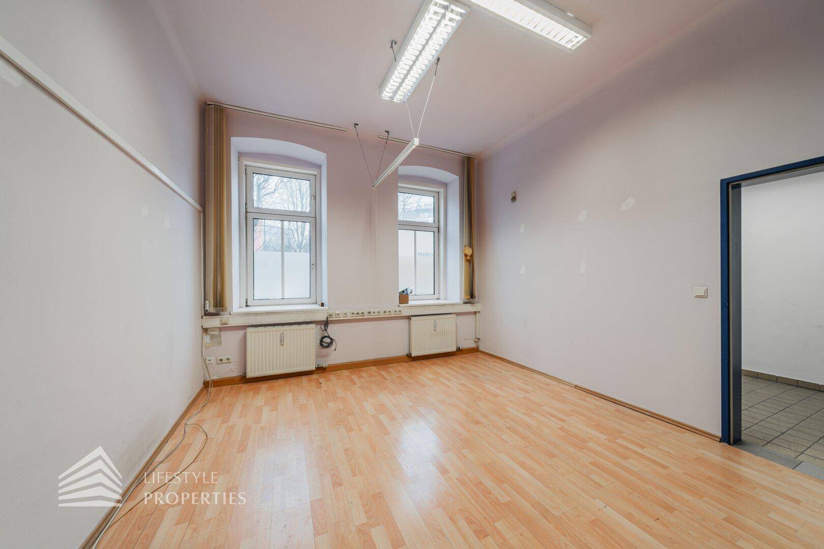 Geräumiges Büro mit Holzboden und zwei Fenstern, die viel Tageslicht hereinlassen, ideal für Arbeitsbereiche.