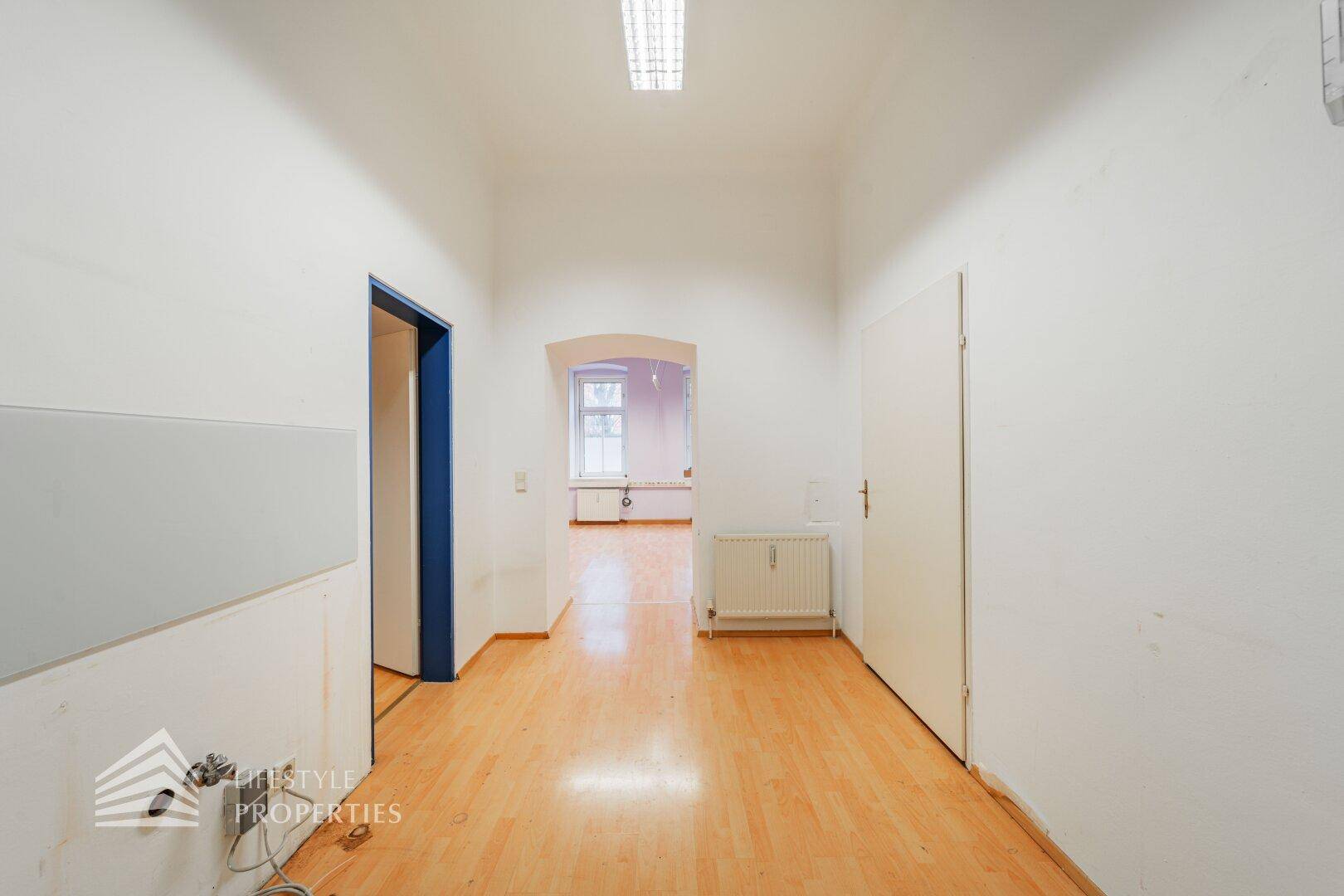 Heller Flur mit Holzboden und weiß gestrichenen Wänden, der einen Blick in einen weiteren Raum erlaubt.