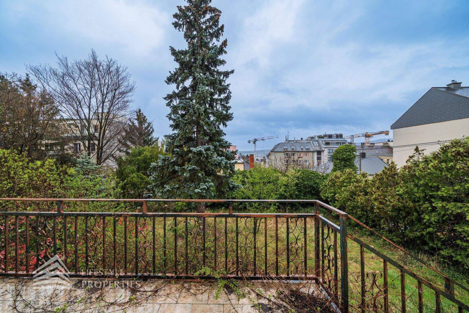 Blick von einem Balkon mit Metallgeländer auf bewachsene Bäume und die Stadt in der Ferne.