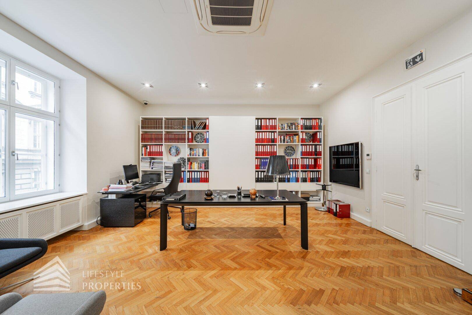 Geräumiges Büro mit Holzparkett, integrierten Regalen voller Aktenordner und einem großen Schreibtisch.