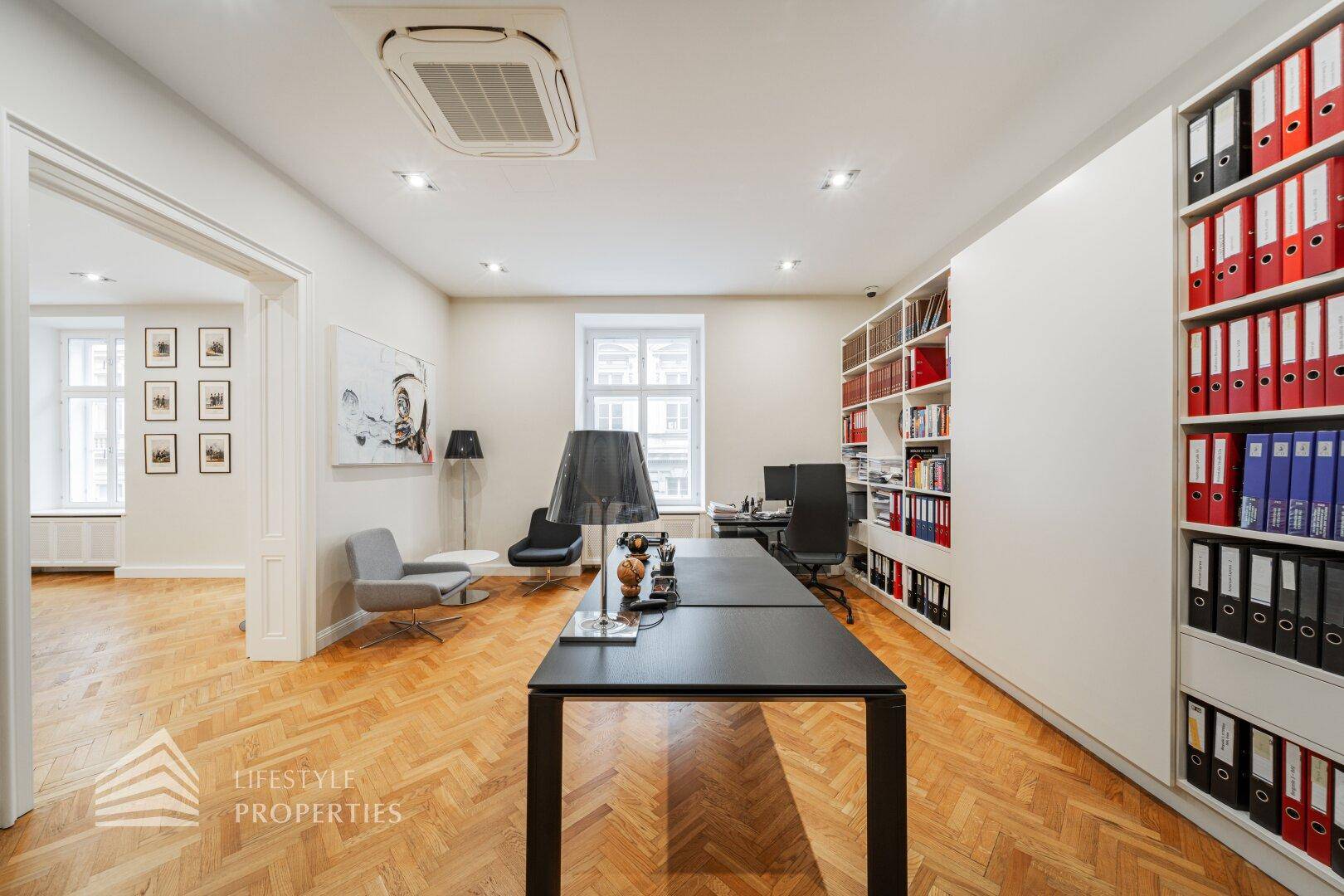 Helles Büro mit Fischgrätparkett, einem großen Schreibtisch und Blick in angrenzende Räume.