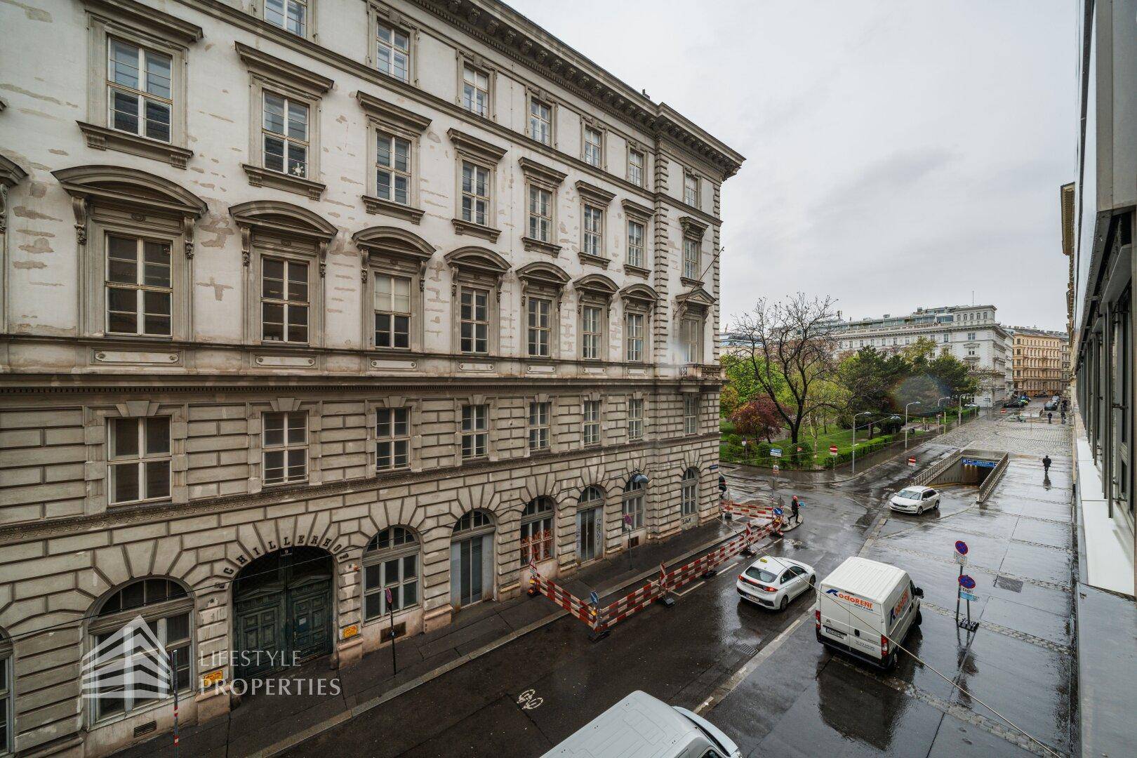 Blick von oben auf eine historische Straße mit Gebäuden, parkenden Autos und einem Park in der Ferne.