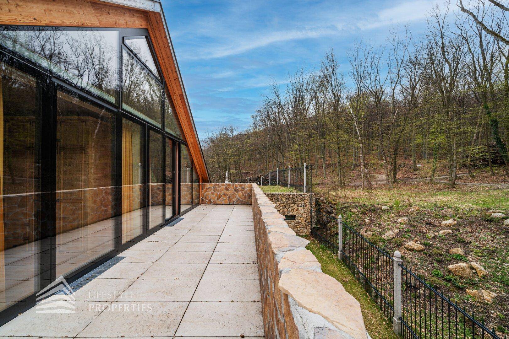 Lange Terrasse entlang des Hauses mit Steinmauer und bodentiefen Fenstern zur Natur.