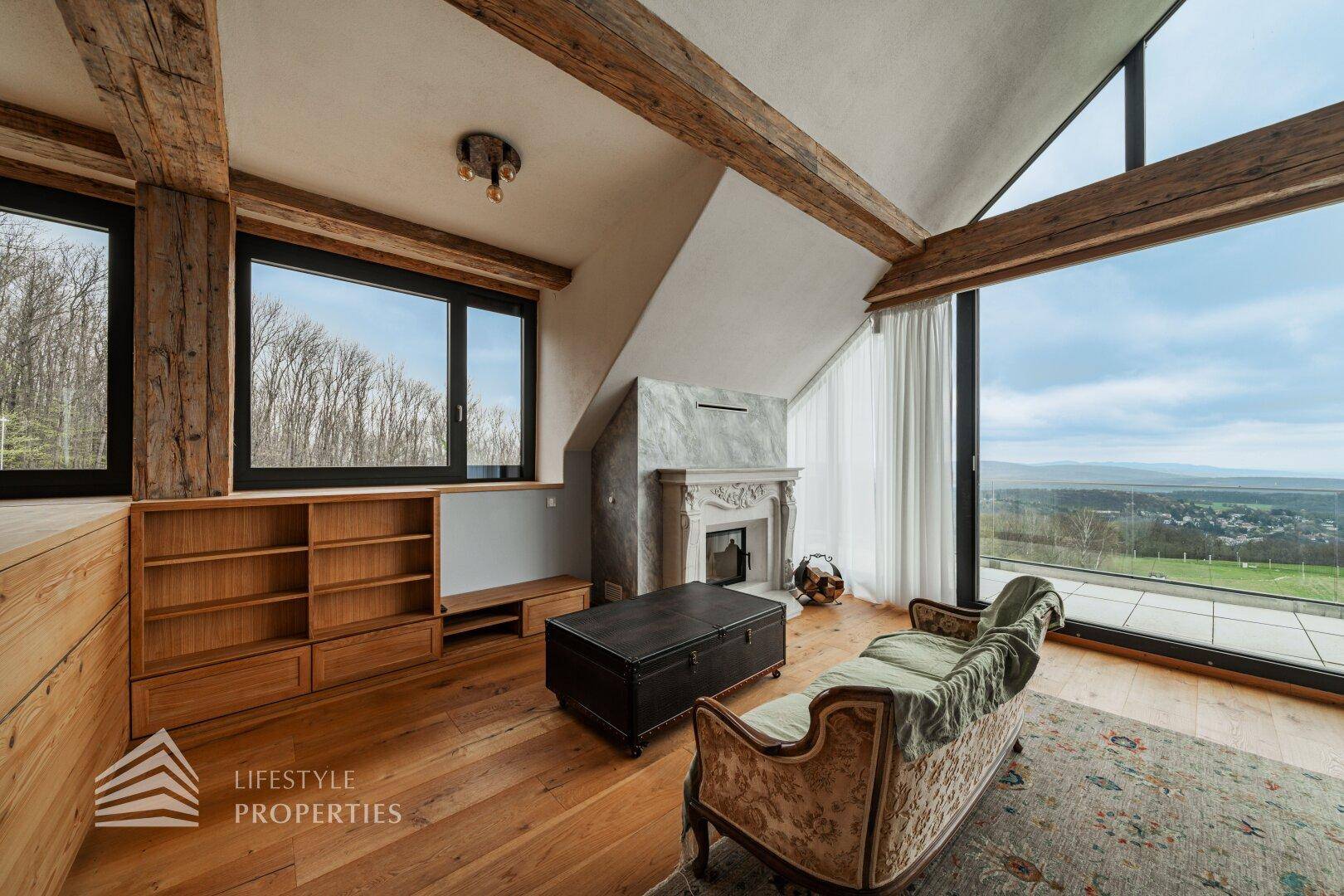 Helles Schlafzimmer mit Kamin, Sofa und großen Fenstern mit Blick auf die bewaldete Umgebung.