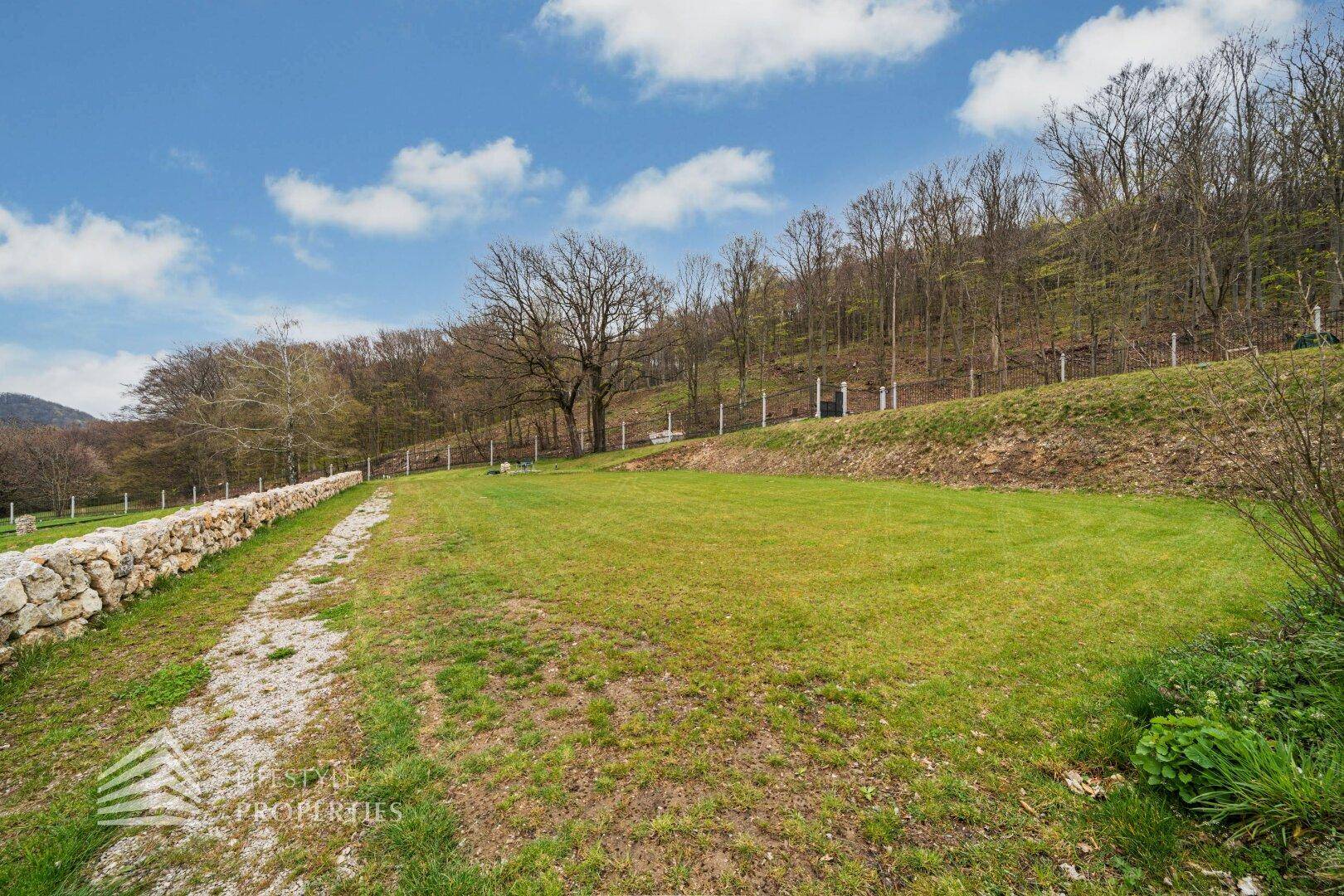 Weitläufige Rasenfläche mit alter Steinmauer und bewaldeten Hügeln im Hintergrund.