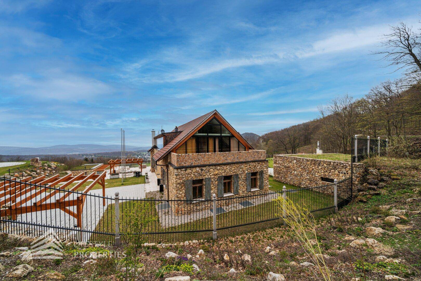 Ansicht des Hauses mit Steinfassade, traditionellem Dach und umzäuntem Grundstück in der Natur.