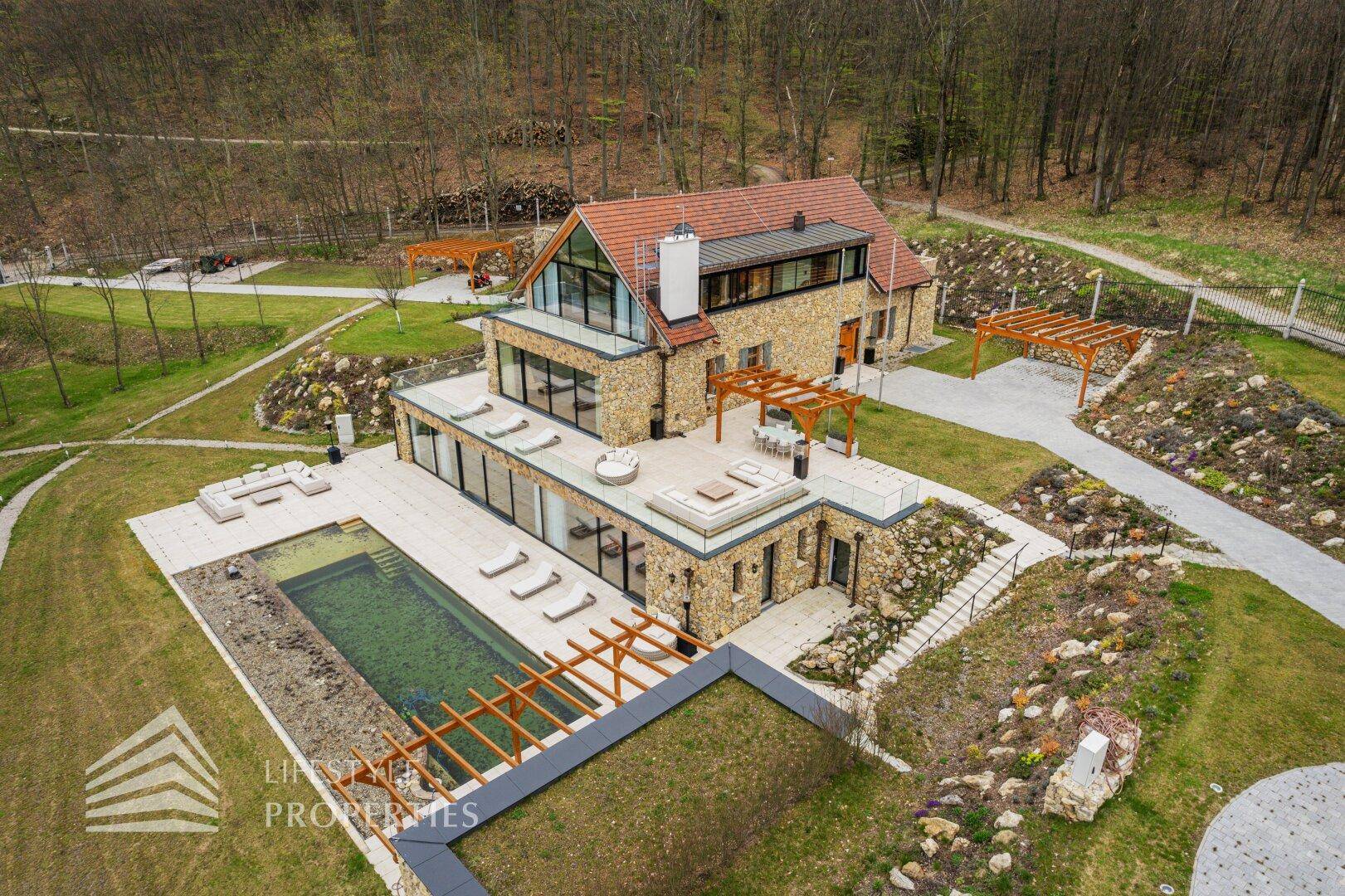 Panoramablick auf das moderne Anwesen mit großem Poolbereich und Terrassen in grüner Hügellandschaft.