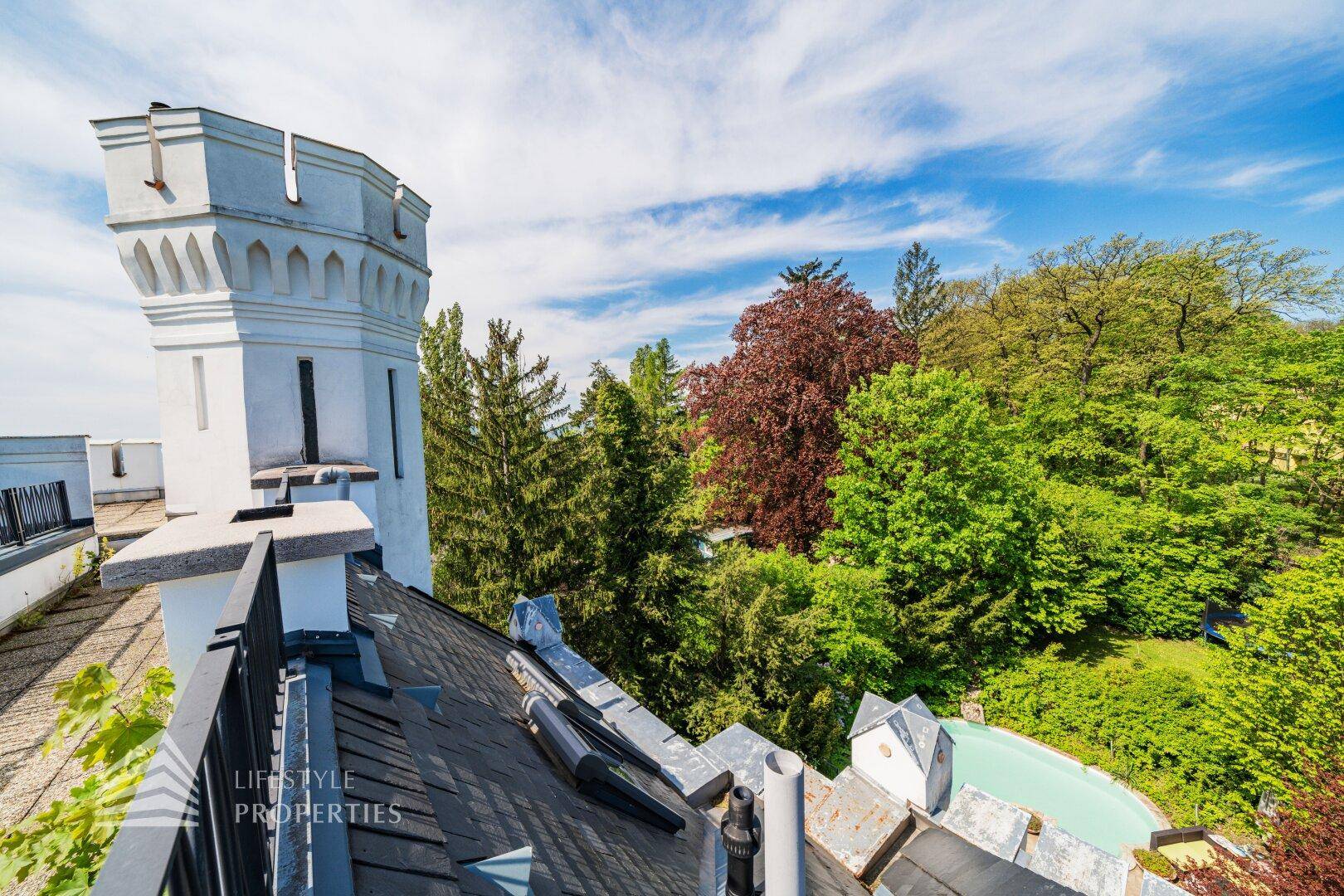 Historisches Gebäude mit markantem Turm, umgeben von üppigem Grün und Bäumen.