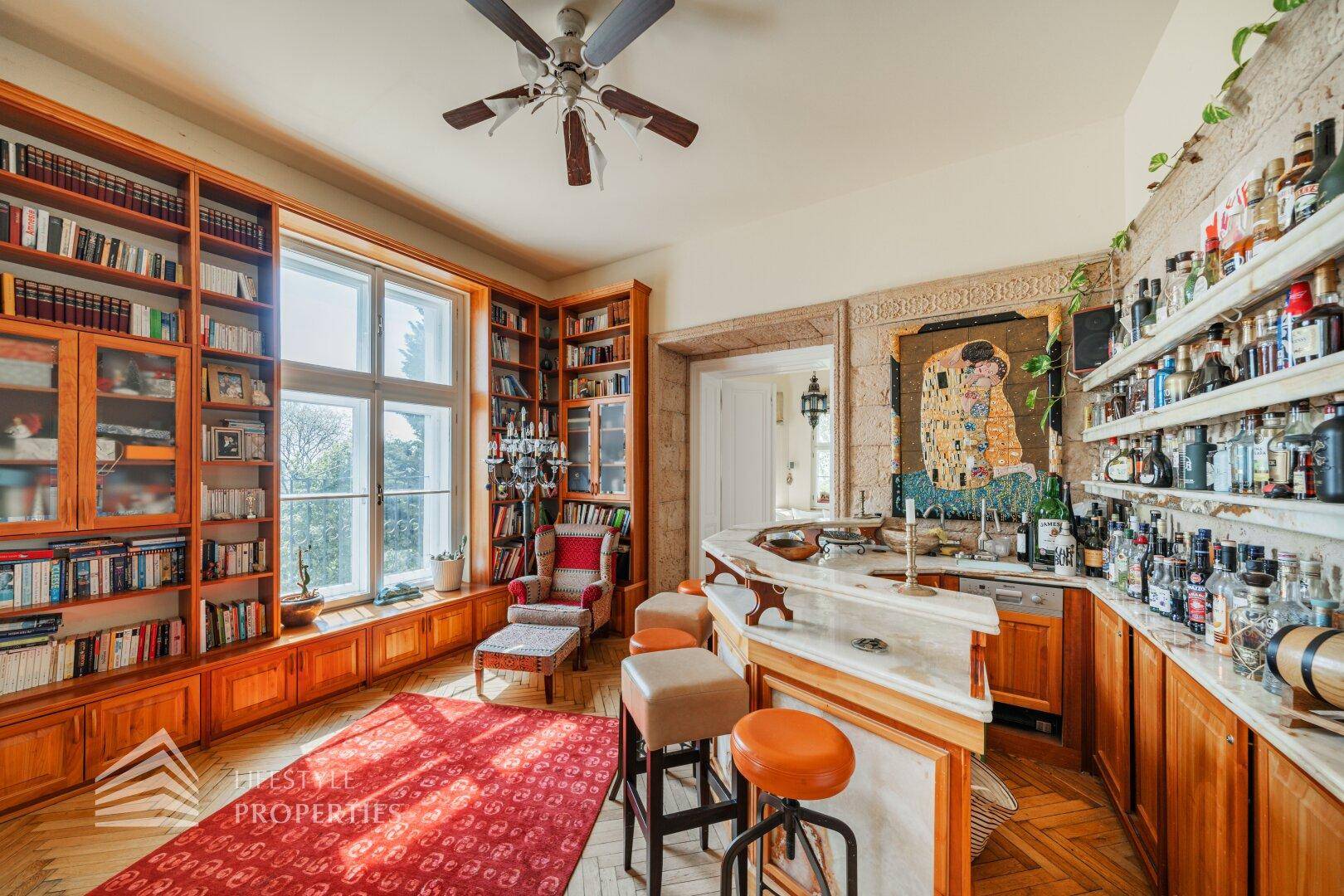 Gemütlicher Barbereich mit umfangreichen Bücherregalen, großen Fenstern und einem Deckenventilator, perfekt zum Verweilen.