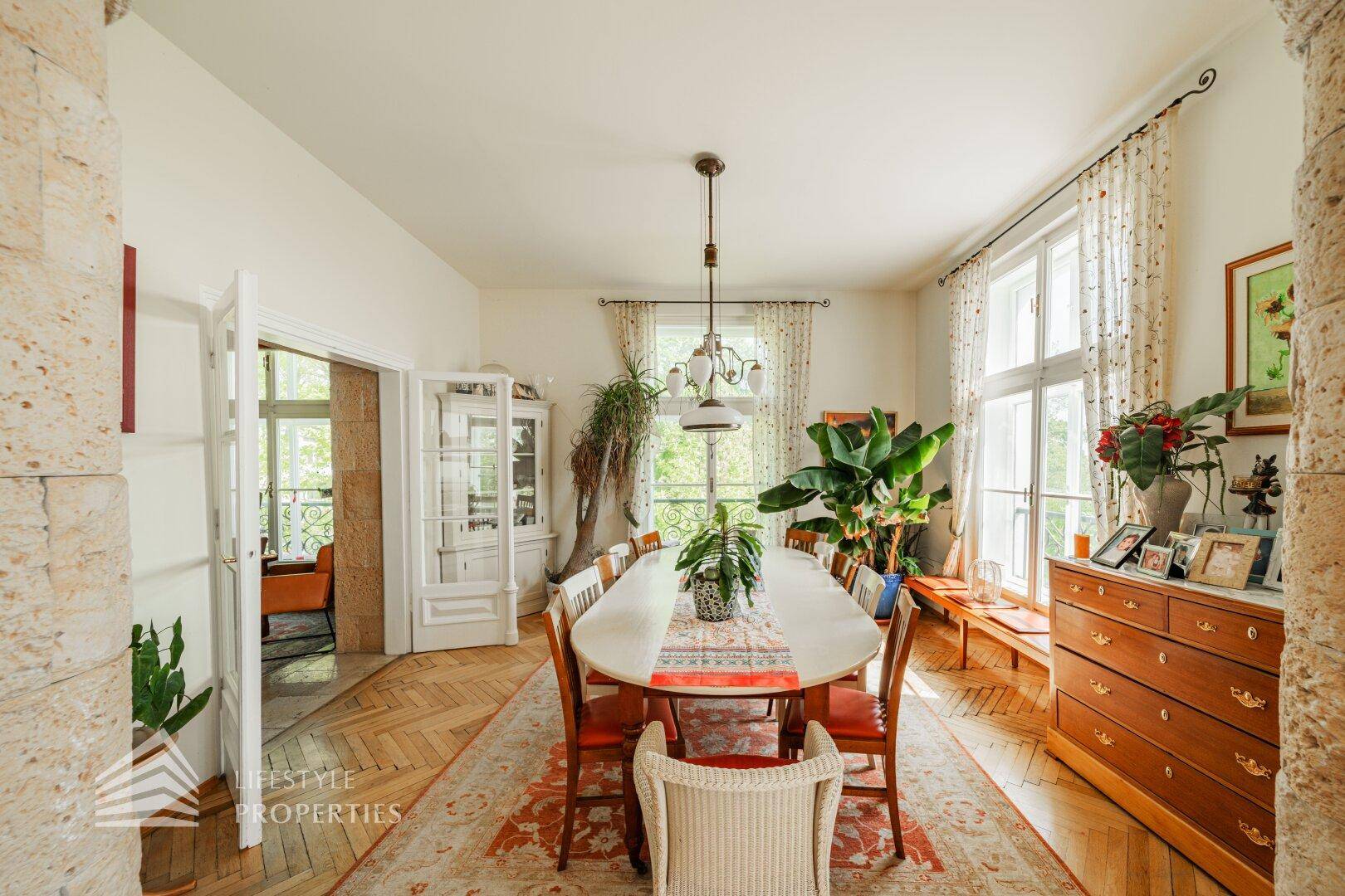 Eleganter Essbereich mit großem ovalen Tisch, hohen Fenstern und üppigen Pflanzen, verbunden mit dem Wohnzimmer.