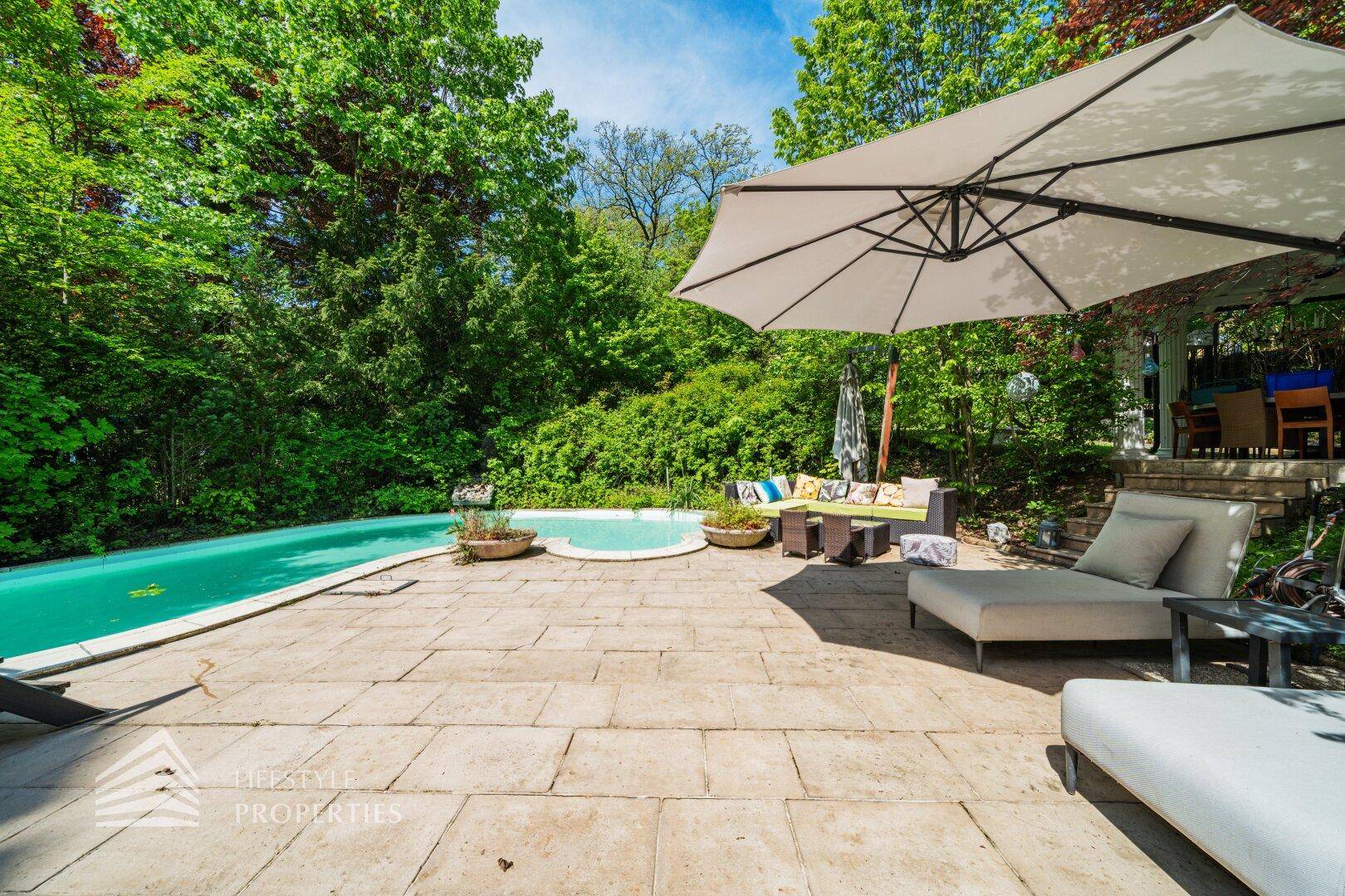 Großzügiger Gartenbereich mit Pool, Liegestühlen und Sonnenschirm, umgeben von Bäumen.