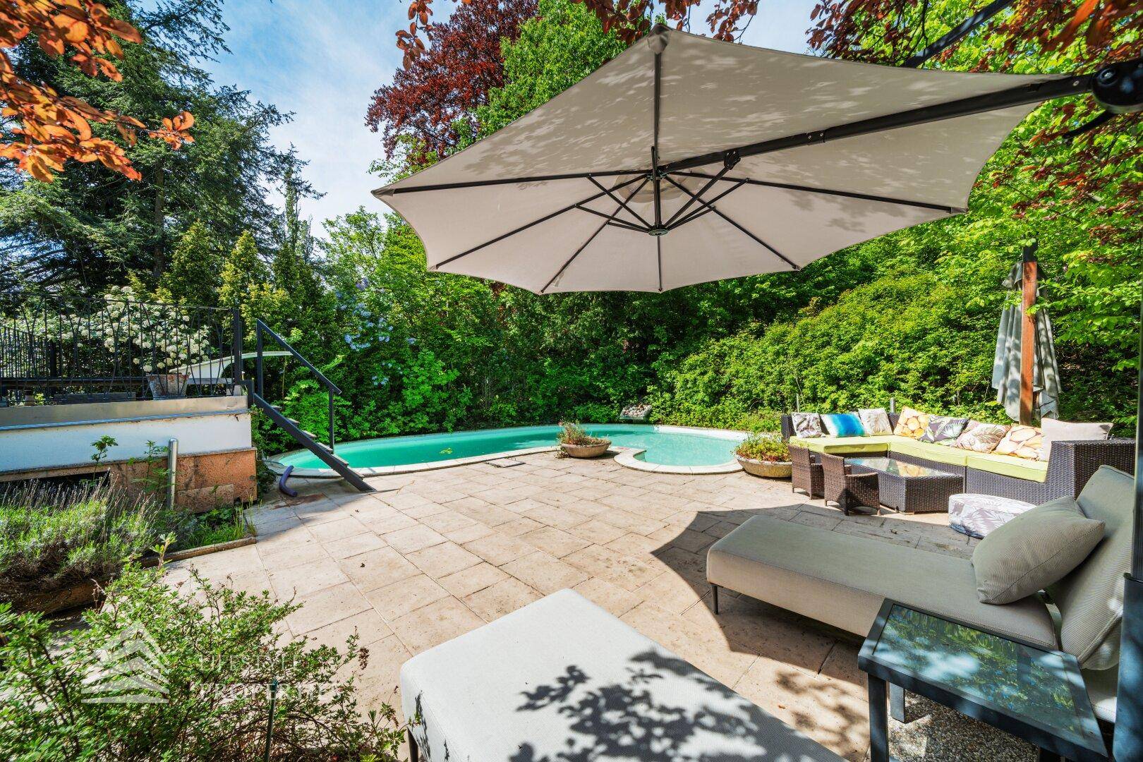 Entspannender Gartenbereich mit Pool, Liegestühlen und großem Sonnenschirm unter Bäumen.