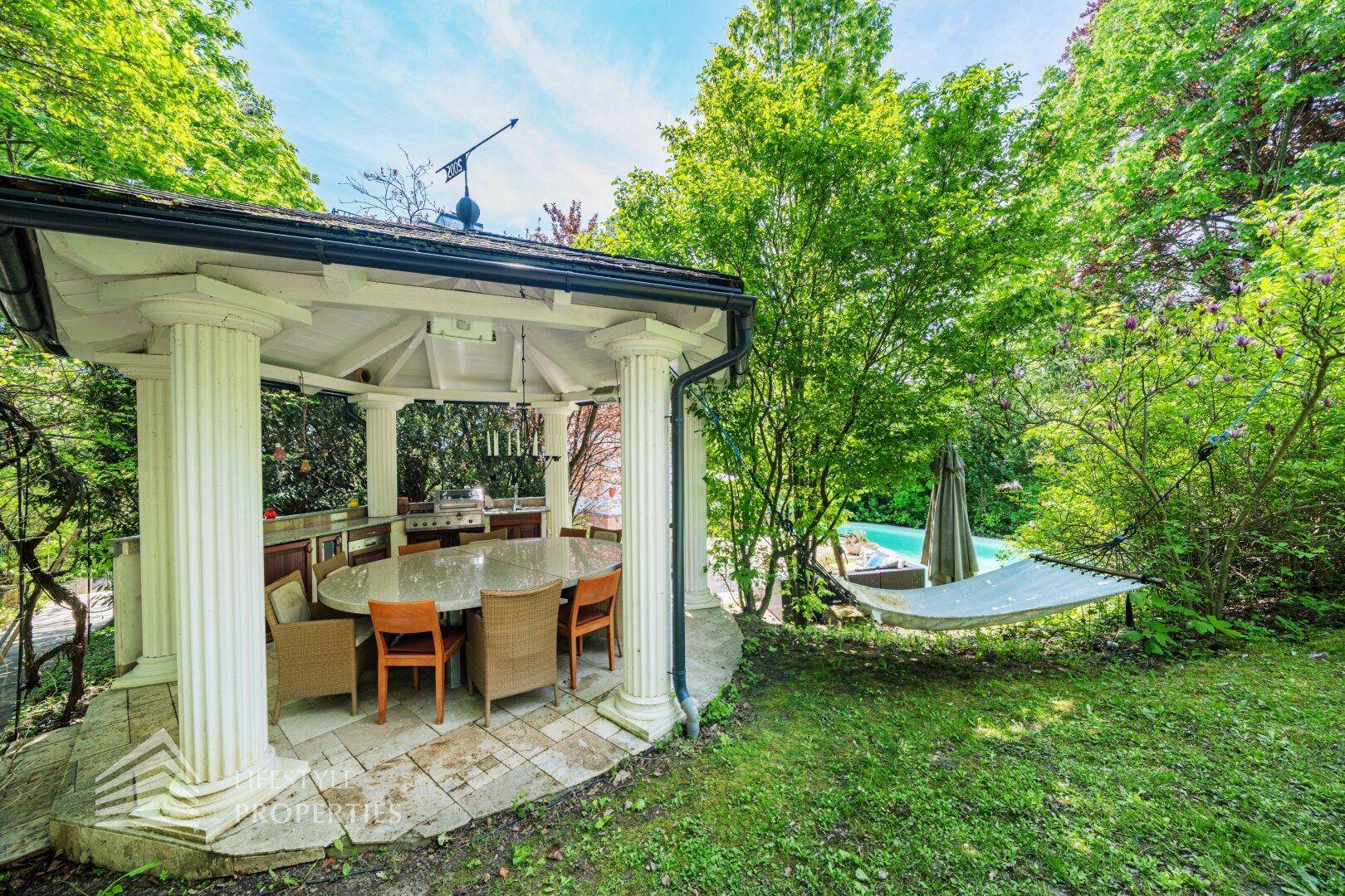 Gemütlicher Gartenpavillon mit Esstisch und Hängematte, umgeben von üppigem Grün.