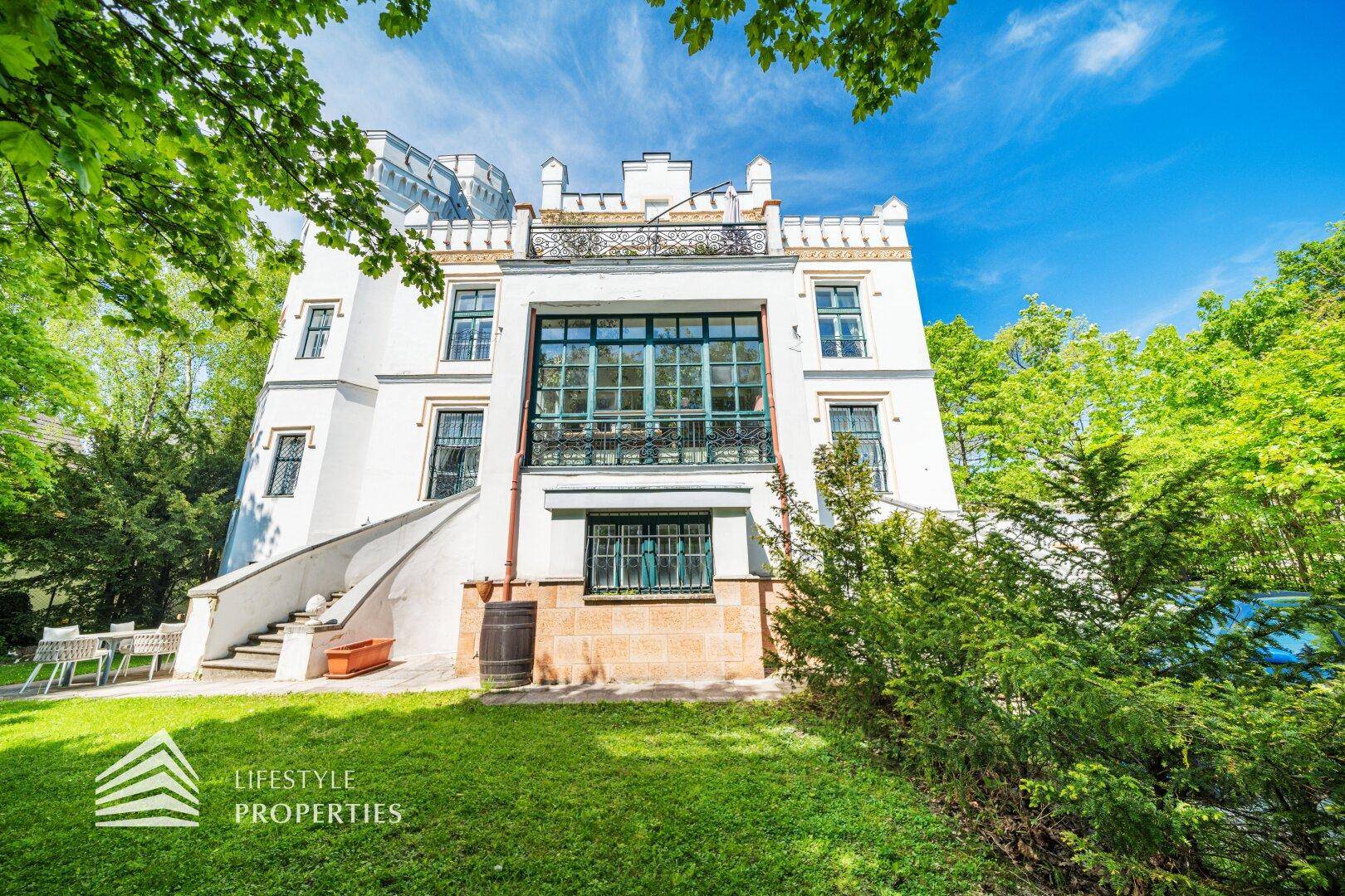 Elegante Fassade des historischen Gebäudes mit großer Glasfront und Balkon, umgeben von Rasenflächen.