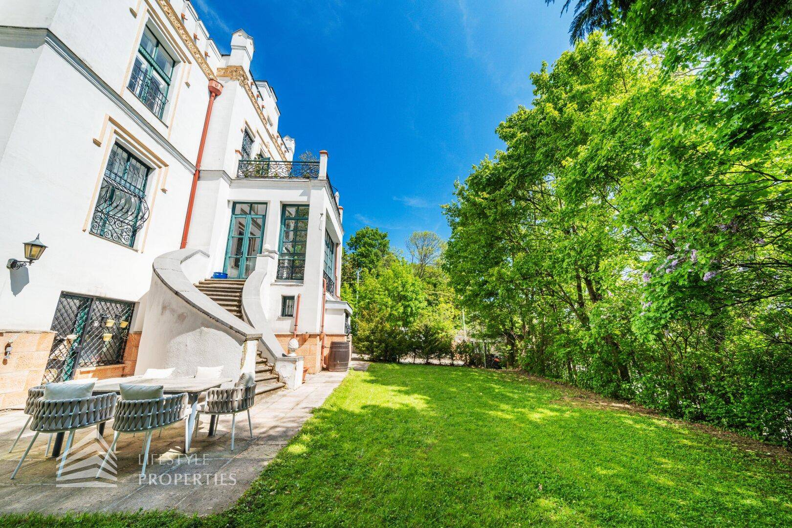 Seitliche Terrasse mit Gartenmöbeln und weitläufiger Rasenfläche, umgeben von hohen Bäumen.