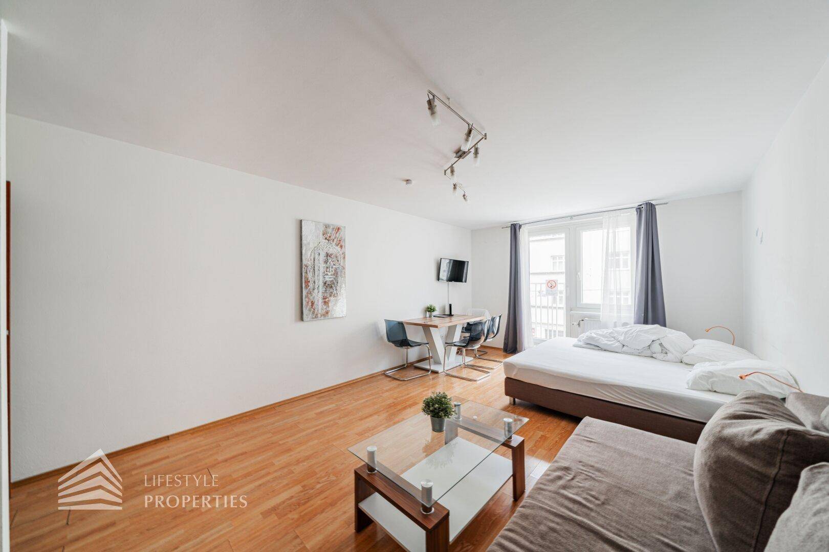Offener Wohnbereich mit Schlafgelegenheit, Esstisch und Zugang zu einem französischen Balkon.