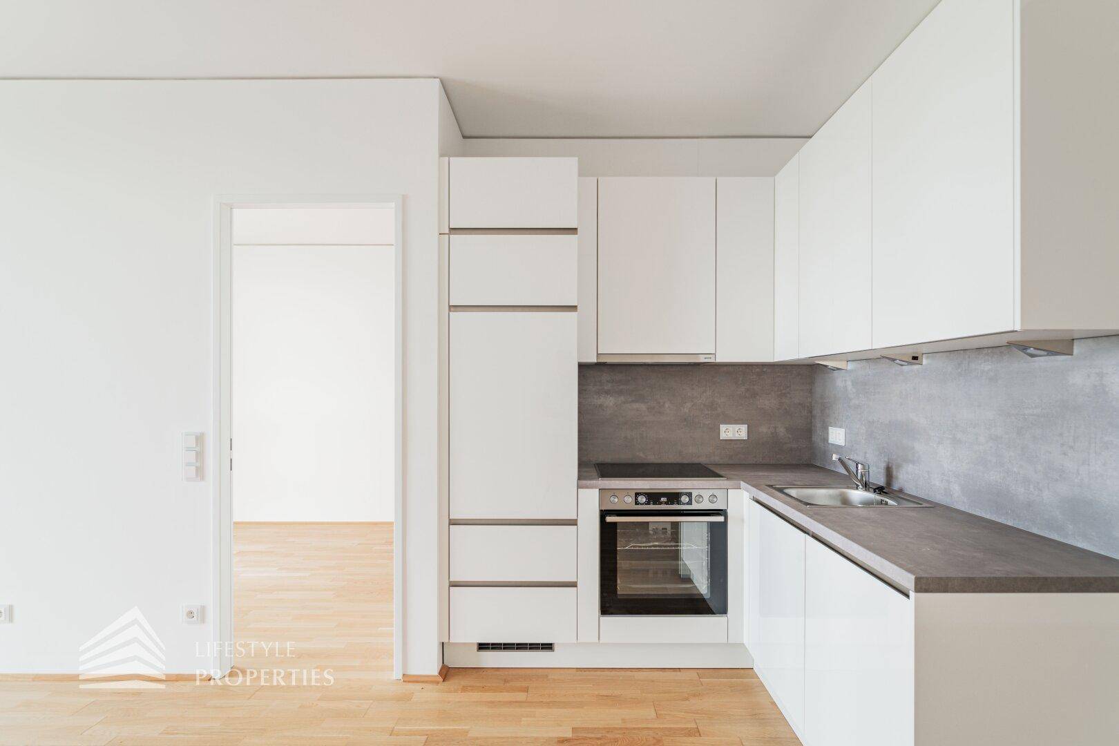 Moderne Einbauküche mit weißen Schränken, integriertem Backofen und grauer Arbeitsplatte.