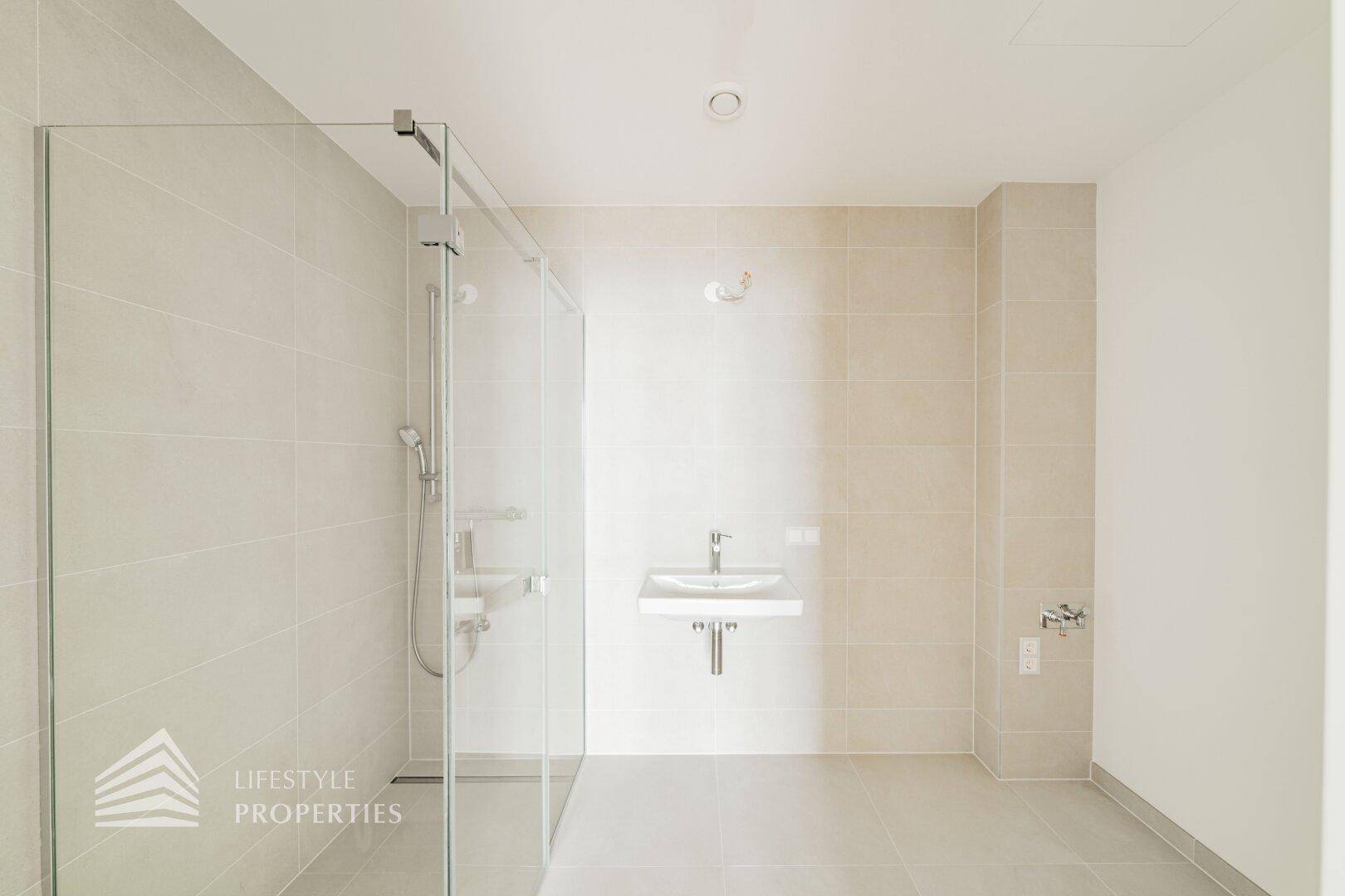 Modernes Badezimmer mit großer Glasdusche, hellem Fliesenboden und wandhängendem Waschbecken.