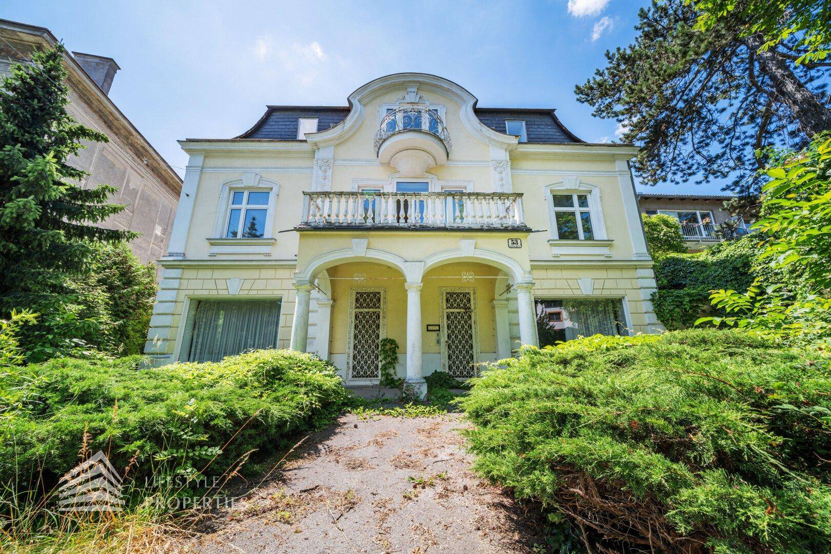 Historische Villa mit gelber Fassade, Säulen und Balkon, umgeben von üppigem Grün.