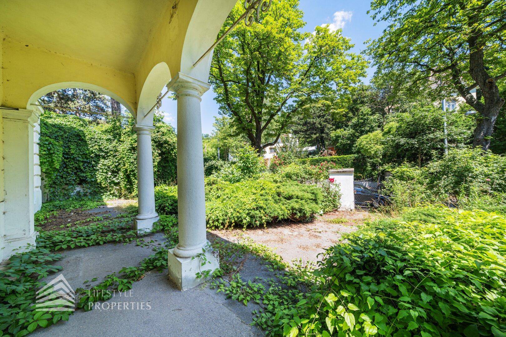 Blick von der überdachten Veranda in den verwilderten Gartenbereich mit alten Bäumen.