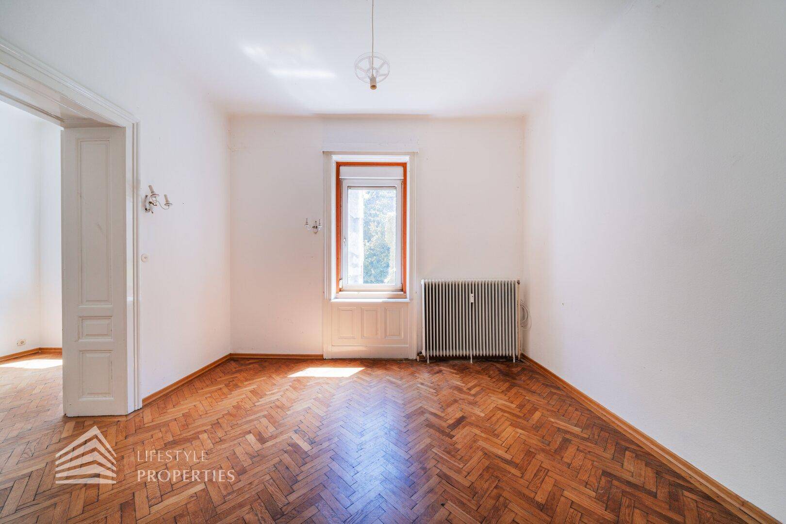 Geräumiges Zimmer mit Fischgrätparkett und großem Fenster, bereit zur Renovierung.