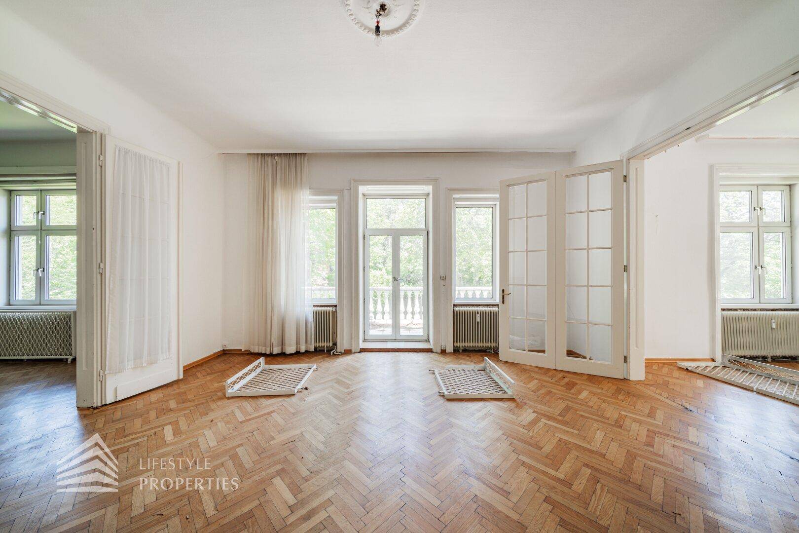 Helles Wohnzimmer mit Fischgrätparkett, hohen Fenstern und Zugang zu einem Balkon.