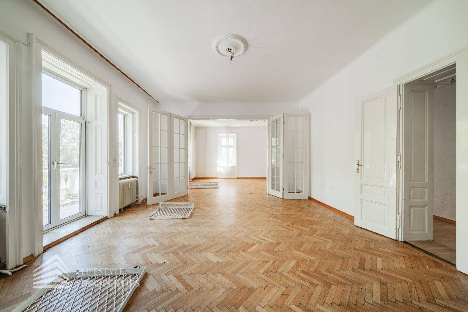 Großzügiger Raum mit Fischgrätparkett, hohen Fenstern und Flügeltüren.