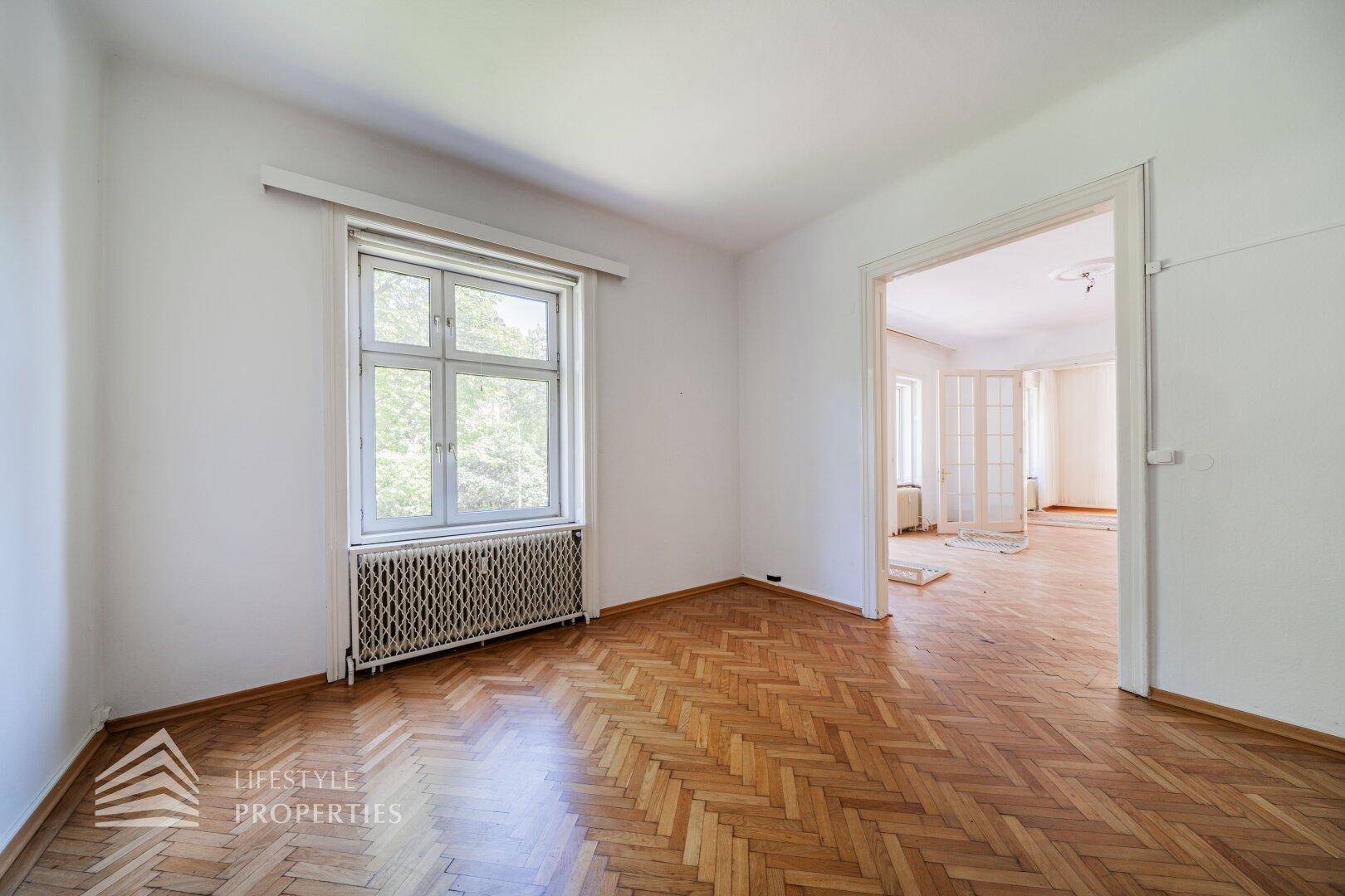 Geräumiges Zimmer mit Fischgrätparkett und Blick ins Grüne durch das Fenster.