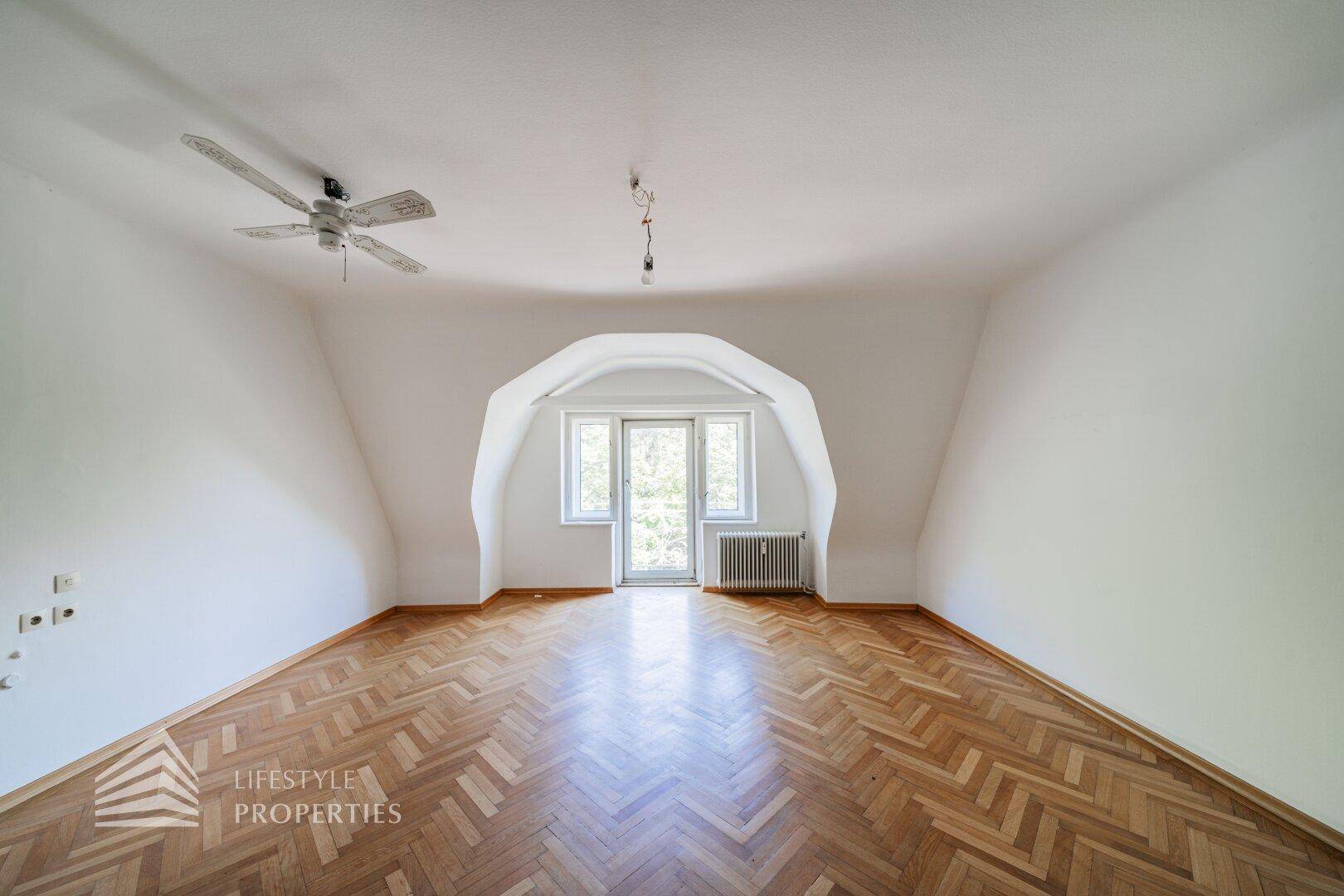 Dachzimmer mit gewölbter Decke, Parkettboden und Fenstertür zum Balkon.