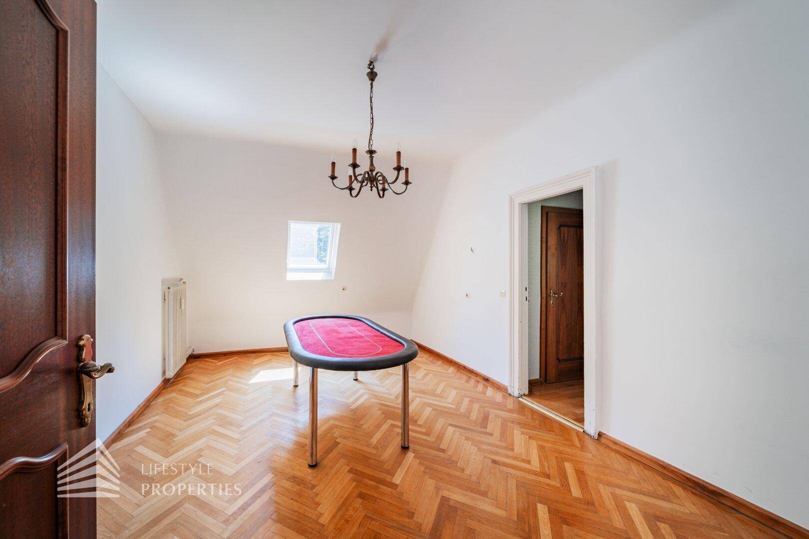 Dachzimmer mit Parkettboden, Dachfenster und einem Spieltisch in der Mitte.