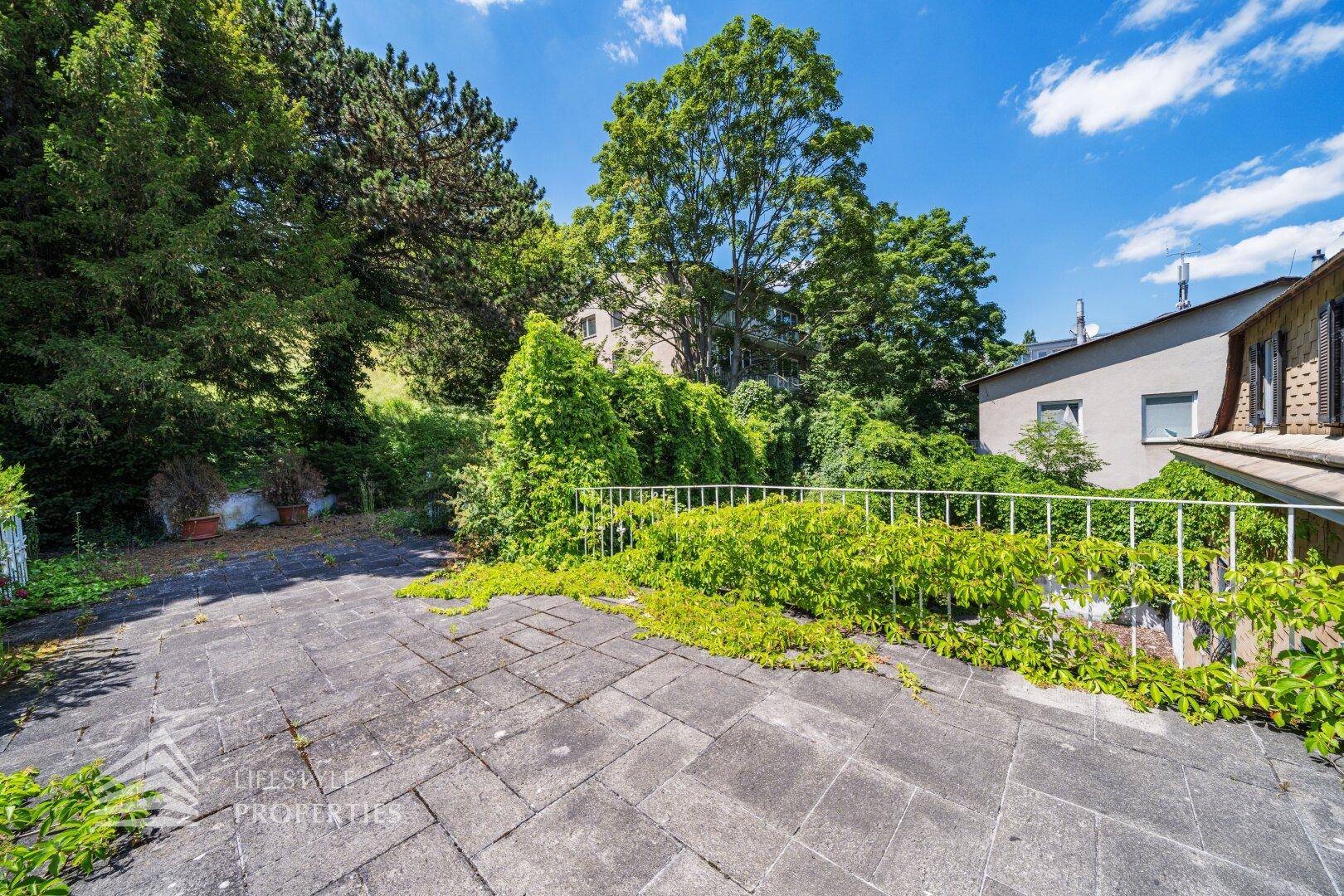 Große gepflasterte Terrasse mit üppiger Begrünung und Blick auf die Umgebung.