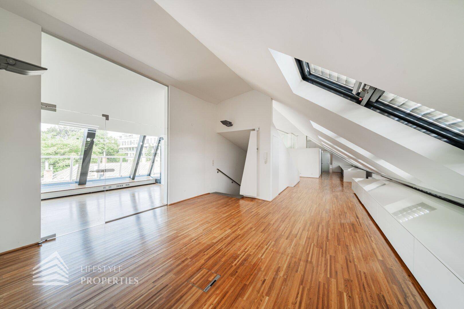 Geräumiger Dachgeschossraum mit Holzboden, schrägen Decken und einer modernen Schiebetür aus Glas.
