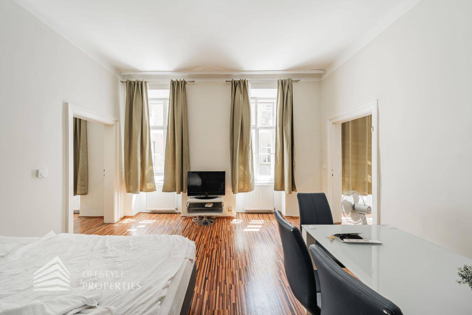 Geräumiges Zimmer mit Holzboden, hohen Fenstern und viel Tageslicht, kombiniert Wohn- und Schlafbereich.