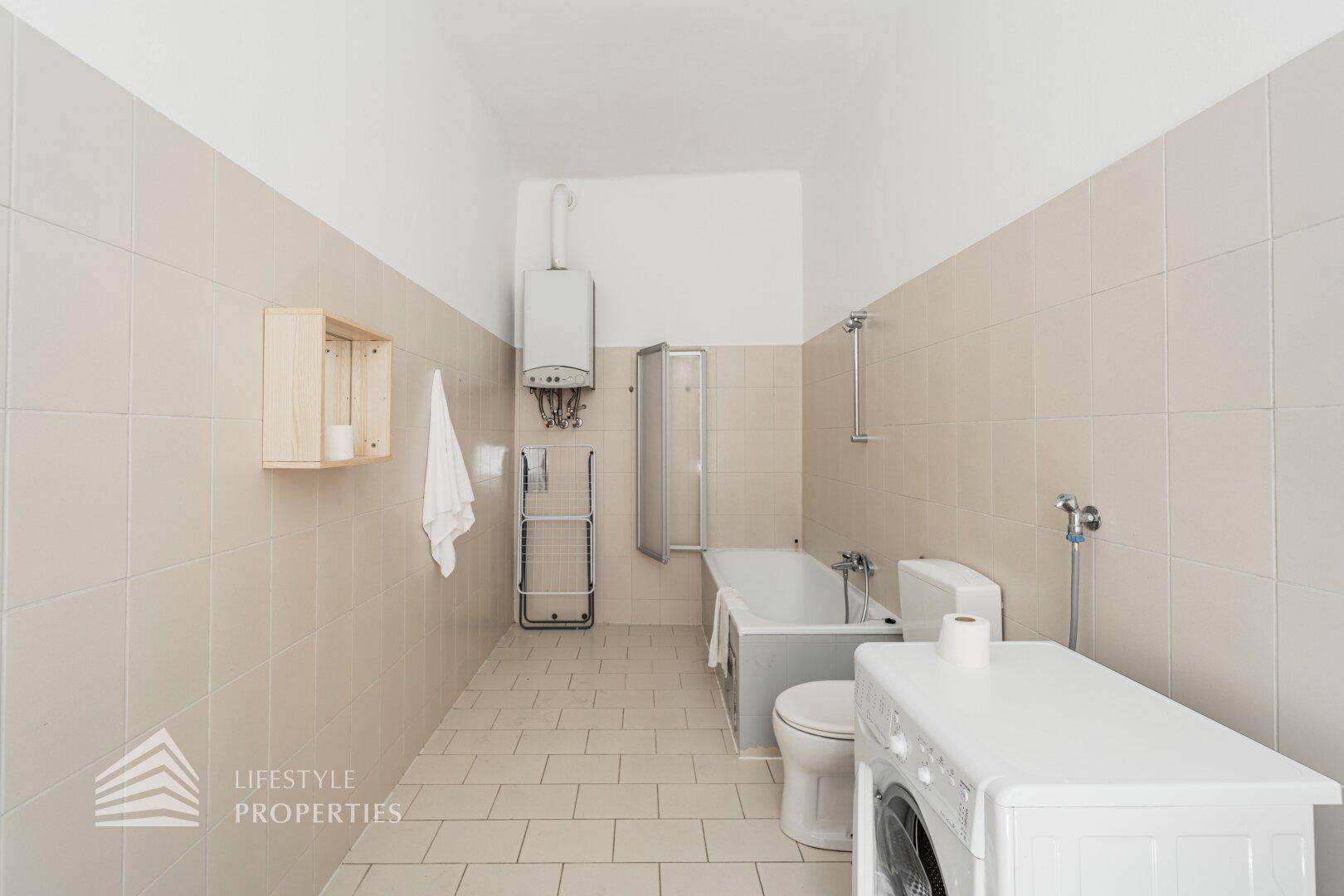 Geräumiges Badezimmer mit Badewanne, WC und Waschmaschine, inklusive Warmwasserbereiter an der Wand.
