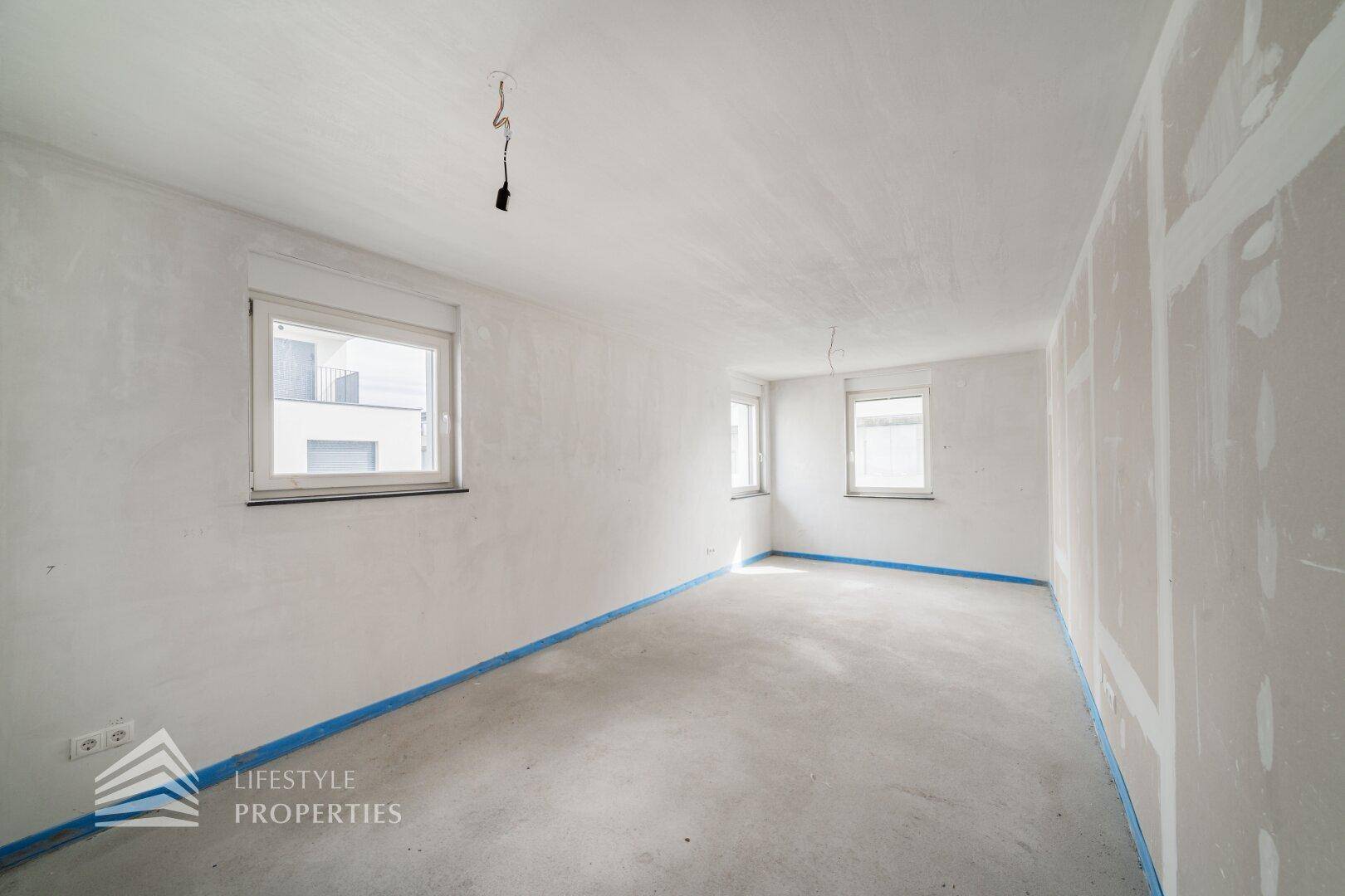 Unfertiges Zimmer mit Rohbetonboden und zwei Fenstern, bereit für den Innenausbau.