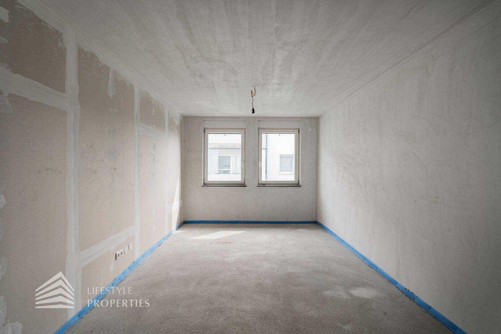 Unfertiges Zimmer mit Rohbetonboden und zwei Fenstern, bereit für den Innenausbau.