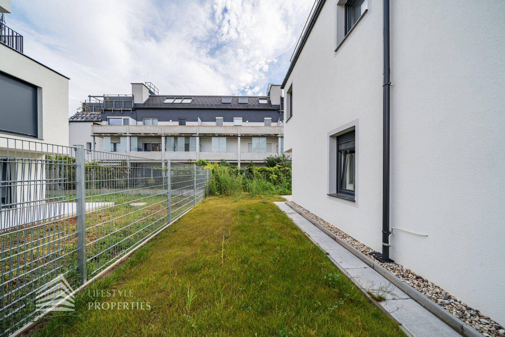 Schmale Rasenfläche entlang der modernen Hausfassade, begrenzt durch einen Metallzaun.