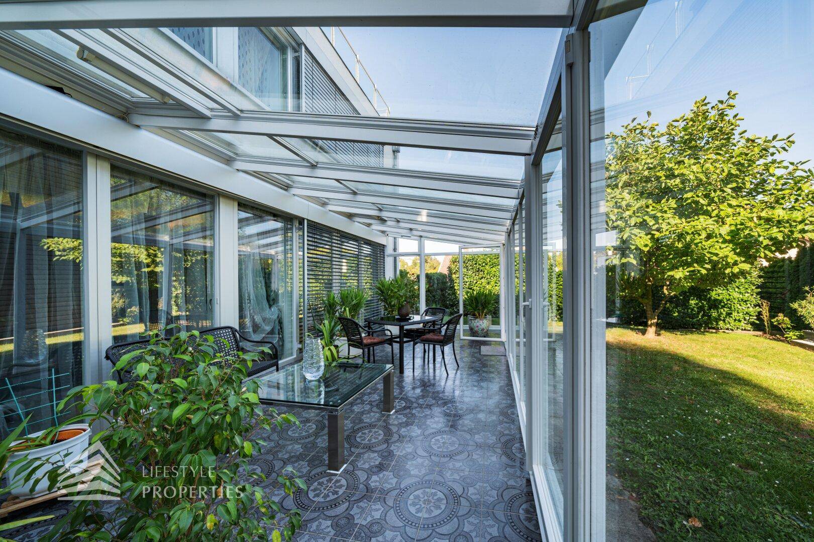 Heller Wintergarten mit Sitzgelegenheiten, Fliesenboden und Blick in den grünen Garten.