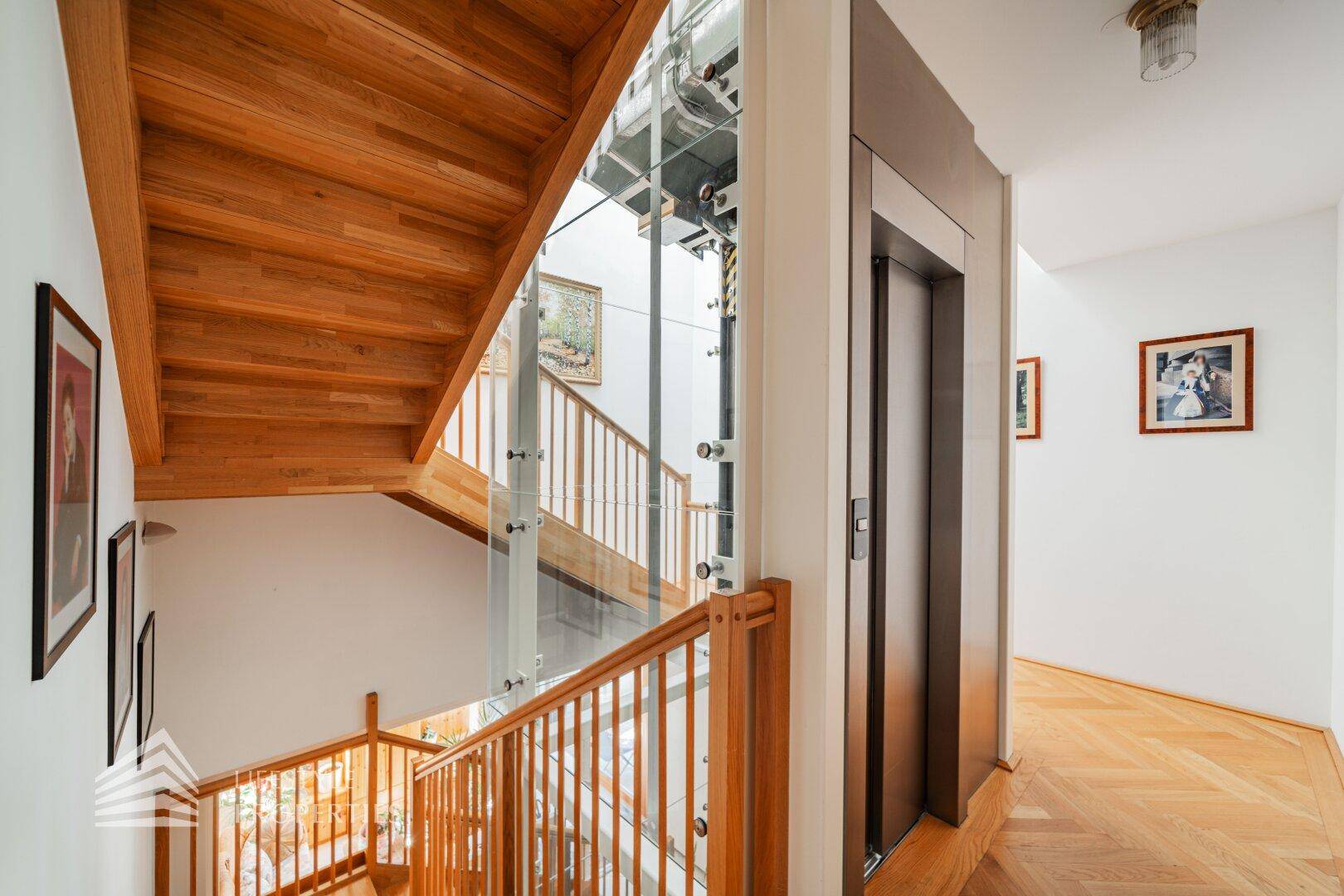 Treppenhaus mit Holzstufen und modernem Glasaufzug, der die Etagen verbindet.