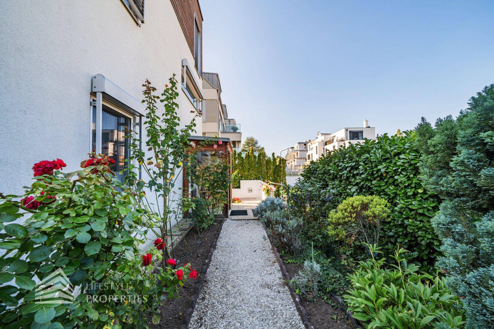 Gepflegter Gartenweg entlang der Hausfassade mit üppiger Bepflanzung und Rosen.