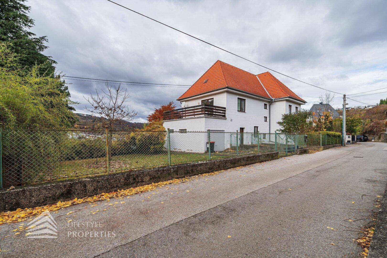 Ansicht des Hauses von der Straße mit Zaun und leichter Hanglage.