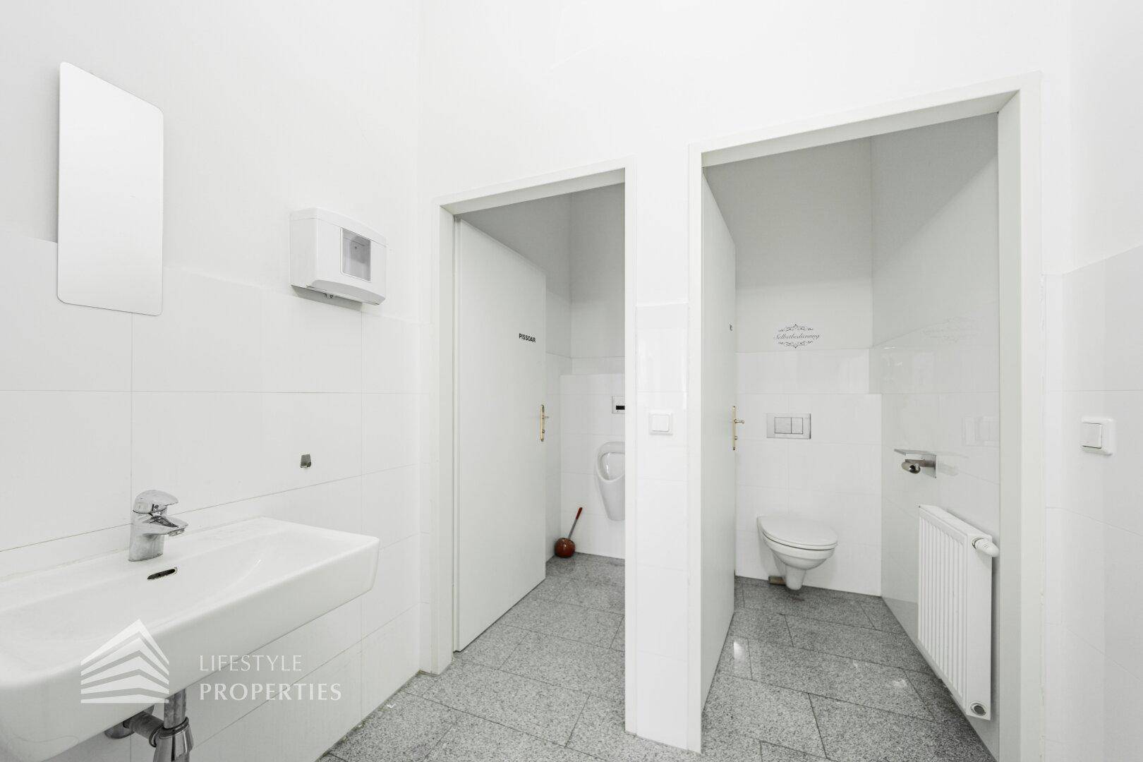 Toilettenbereich mit Waschbecken, Urinal und WC, getrennt durch Türen in einem hellen Raum.