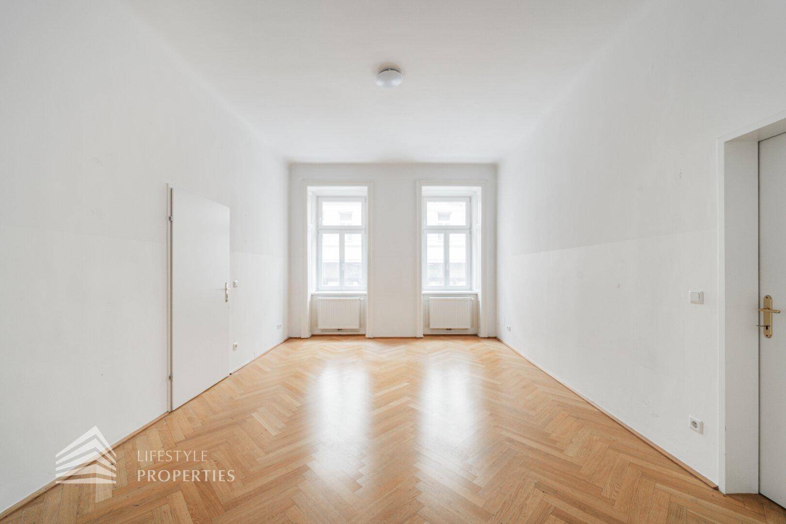 Heller Raum mit Fischgrätparkett und zwei Fenstern, bietet viel Tageslicht und eine ruhige Atmosphäre.